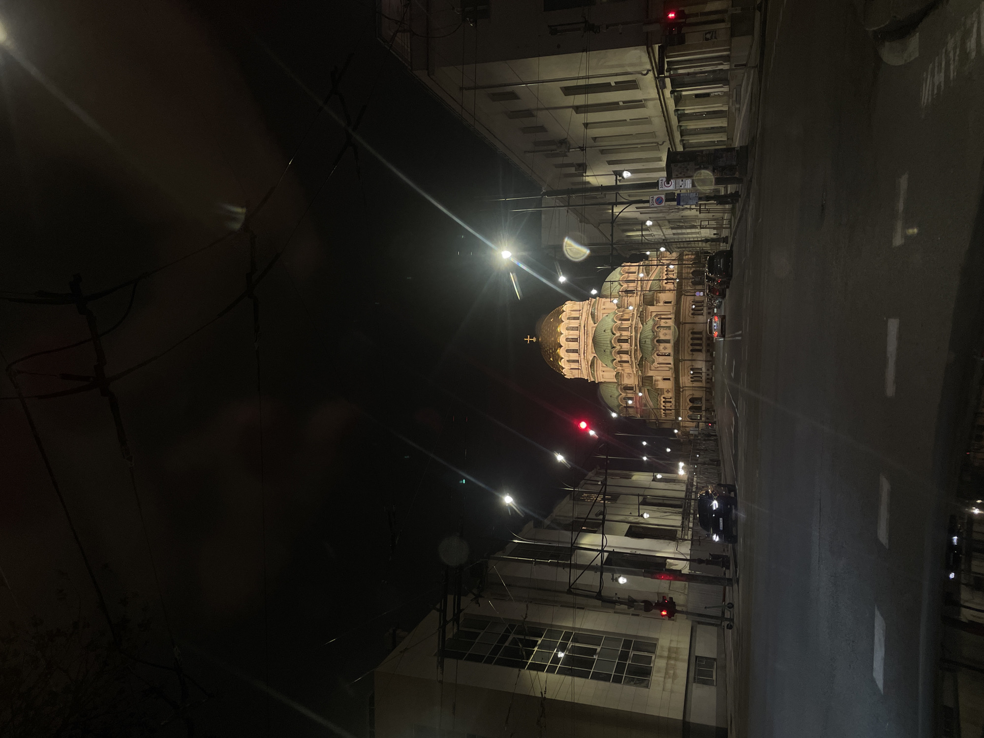 The image depicts a nighttime view of the Alexander Nevsky Cathedral in Sofia, Bulgaria, taken from an urban street. The cathedral, brightly lit, dominates the center of the scene. The street is dark with some street lights illuminating the pavement. Buildings line either side of the street. A car drives down the road. Traffic lights are visible. Utility poles and wires are prominent against the night sky. The overall impression is of a quiet night in Sofia, with the iconic cathedral as the focal point.