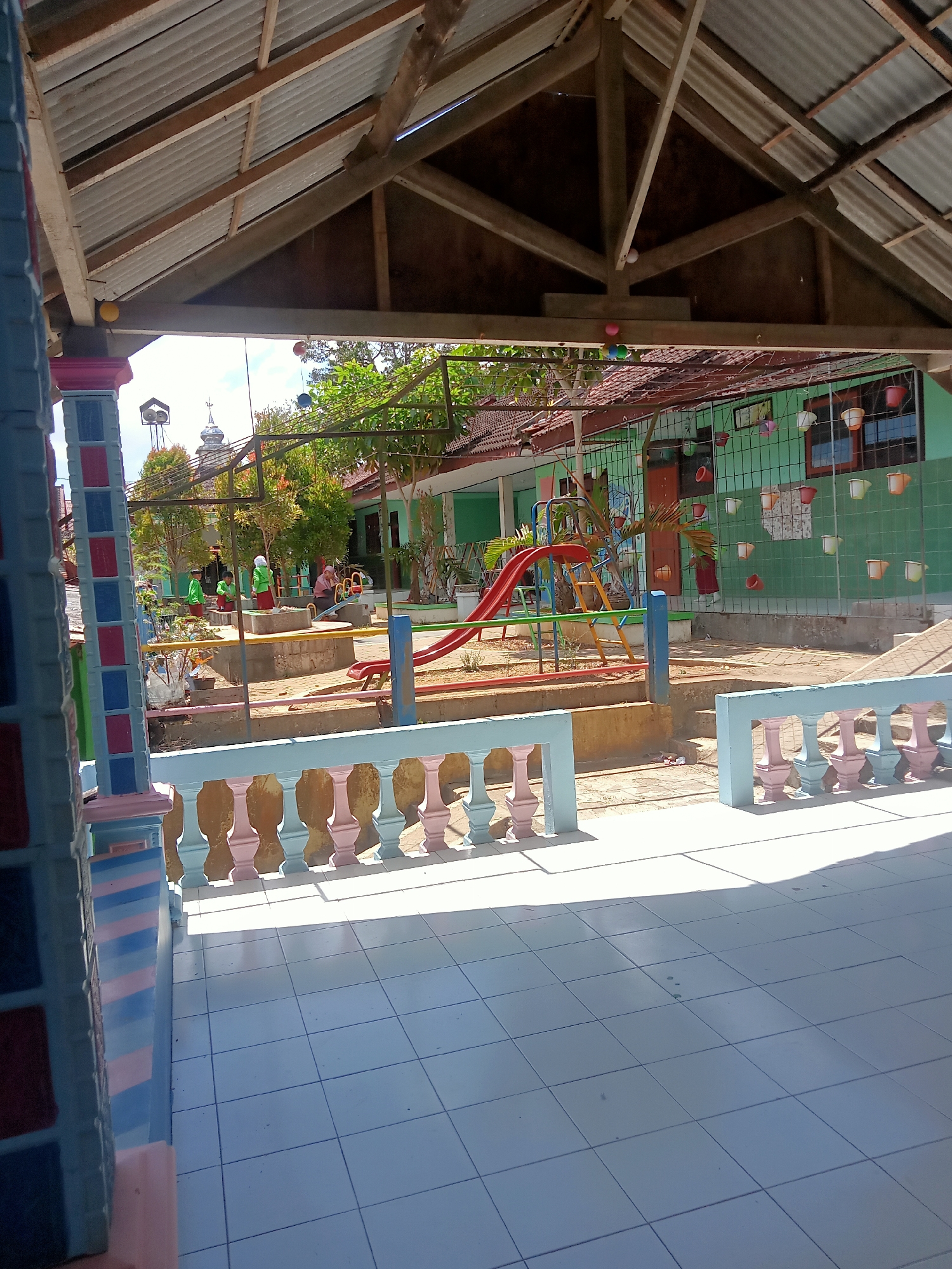 This image captures a view of a playground area at what appears to be a school or community center in Pule, Indonesia. The foreground features a tiled floor under a covered structure with a corrugated metal roof supported by wooden beams. A colorful railing, painted in pastel shades of pink and light blue, separates the tiled area from the playground.
The playground itself includes a red slide with a yellow ladder, and a blue platform. In the middle ground, several children are visible, some wearing green shirts and white headscarves, suggesting a school setting. They are standing near a low wall and a sandpit area. Beyond the playground, a green building with windows and hanging planters is visible. A water tower with a dome-like structure can be seen in the distance, along with trees and a clear sky. The overall scene suggests a bright, sunny day.