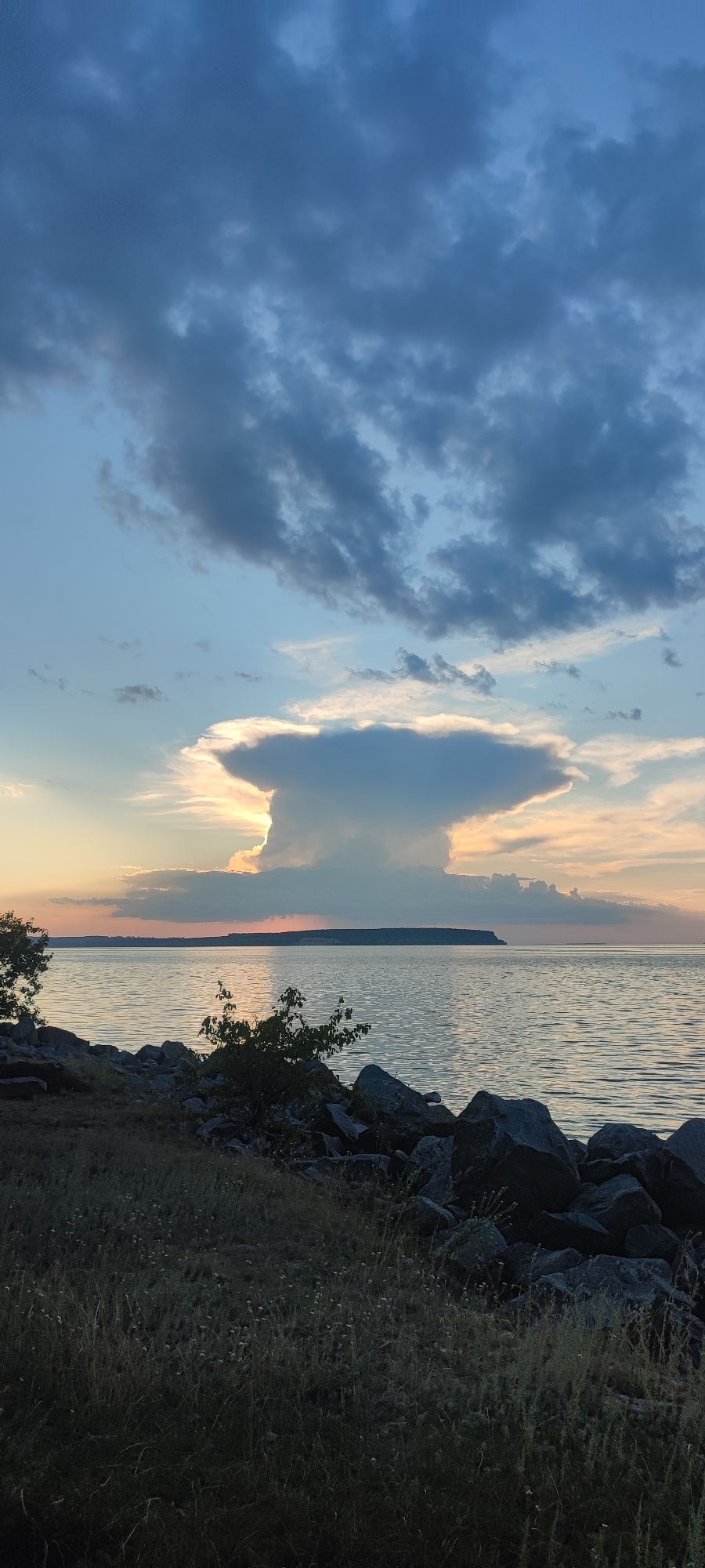 This vertical, full shot captures a serene sunset scene over a body of water. The sky is a canvas of blues and grays, with dramatic, dark clouds congregating at the top, hinting at a recent or impending storm. Below these, lighter, wispy clouds catch the fading sunlight, casting a warm glow and outlining a large, mushroom-shaped cumulonimbus cloud with an incandescent halo. The horizon is a dark, flat line, possibly a distant landmass. The water is calm, reflecting the muted colors of the sky and horizon. In the foreground, a rocky shoreline is visible, covered with dry, wispy grass and a few small, dark green bushes. Large, dark boulders are scattered along the bank, leading the eye towards the water. The overall mood is peaceful and awe-inspiring, with a touch of drama from the imposing cloud formation. The lighting suggests the time of day is late afternoon or early evening, with the sun low on the horizon. There is no visible text in the image.