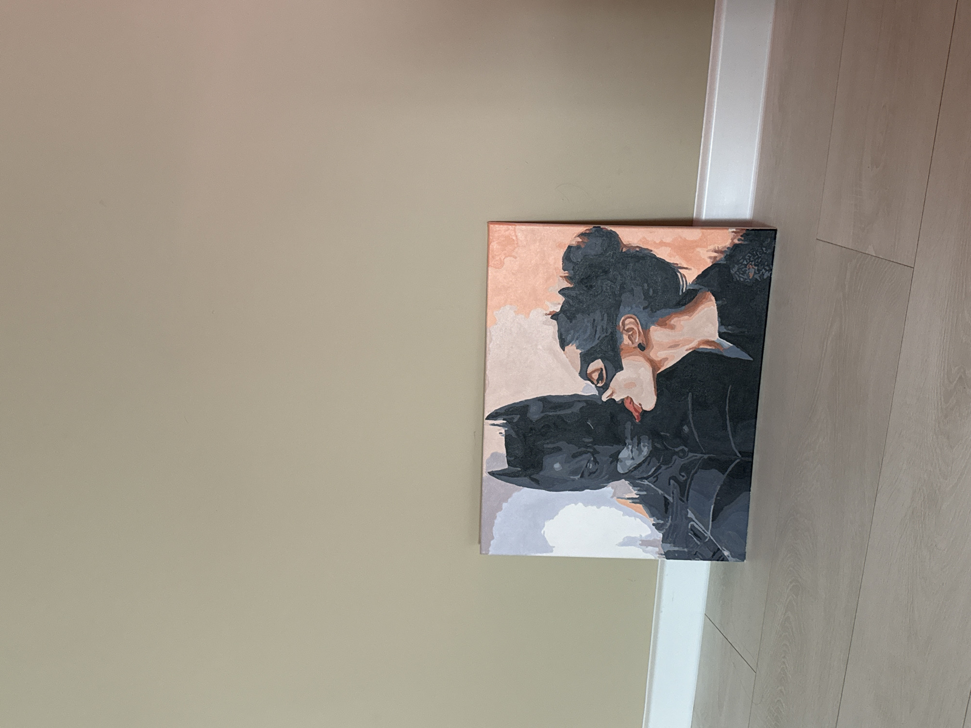 The image displays a vertically oriented painting hanging on a light beige wall, with a white doorframe and light wood laminate flooring visible to the right.
The painting itself features two stylized figures engaged in an intimate moment. The figure on the left is identifiable as Batman, characterized by his dark grey cowl with pointed ears, shown looking downwards. The figure on the right, resembling Catwoman, has light skin, a dark bun hairstyle, and a black mask covering her eyes, with red lips visible. Her head is tilted towards Batman, and their faces are positioned very close, suggesting a tender embrace or a kiss. The artwork uses a block-color aesthetic, predominantly in shades of dark grey, black, various skin tones, against an abstract background of soft peach, light orange, grey, and white washes.
The overall scene suggests an interior setting, likely a home, with the painting serving as decorative art. The wall is plain, and the flooring features distinct planks. The lighting is soft and even. There is no visible text in the image.