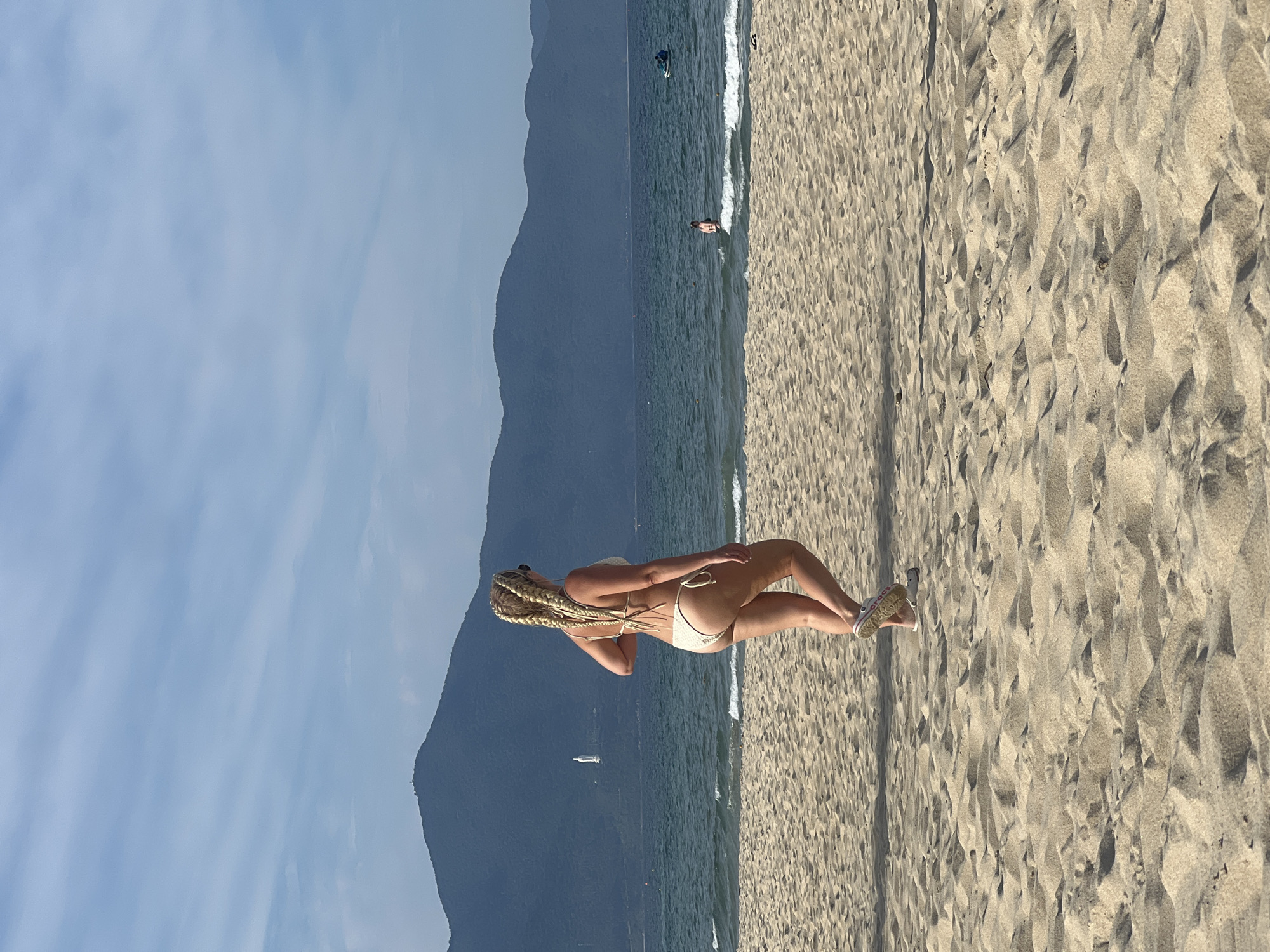 In Đà Nẵng, Vietnam, a woman with blonde braided hair, wearing a cream-colored bikini, walks along a sandy beach. She is viewed from behind, facing towards the ocean and the distant, mountainous coastline. The sea is calm with gentle waves. In the background, a statue of a woman, possibly the Lady Buddha, is visible on a hillside, and further out at sea, a small sailboat can be seen. The sky is a clear blue with wispy white clouds. The sand in the foreground is textured with ripples and shadows, indicating sunlight.
