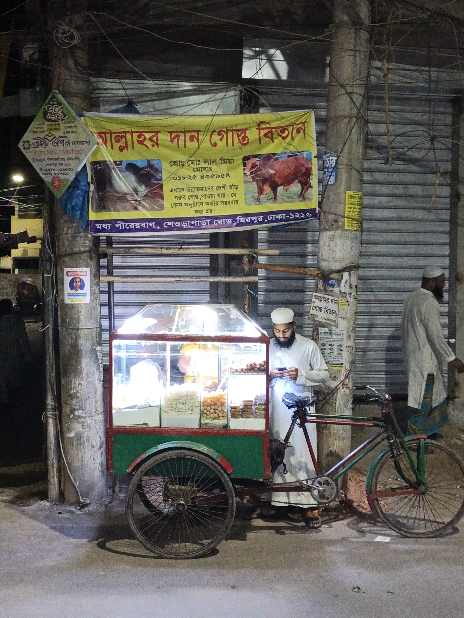 A street scene in Dhaka, Bangladesh, features a man in Muslim attire using a phone near a three-wheeled food cart with a lighted display case containing various snacks. The cart is attached to a bicycle and is adorned with red and green paint. Behind the cart, a sign advertises "Allah's Gift Meat Shop" with images of bulls and text in Bengali, indicating the sale of high-quality beef. Another man in Muslim attire walks past in the background. The setting includes concrete poles covered with posters, advertisements, and electrical wires, with a corrugated metal roll-up door visible in the background.