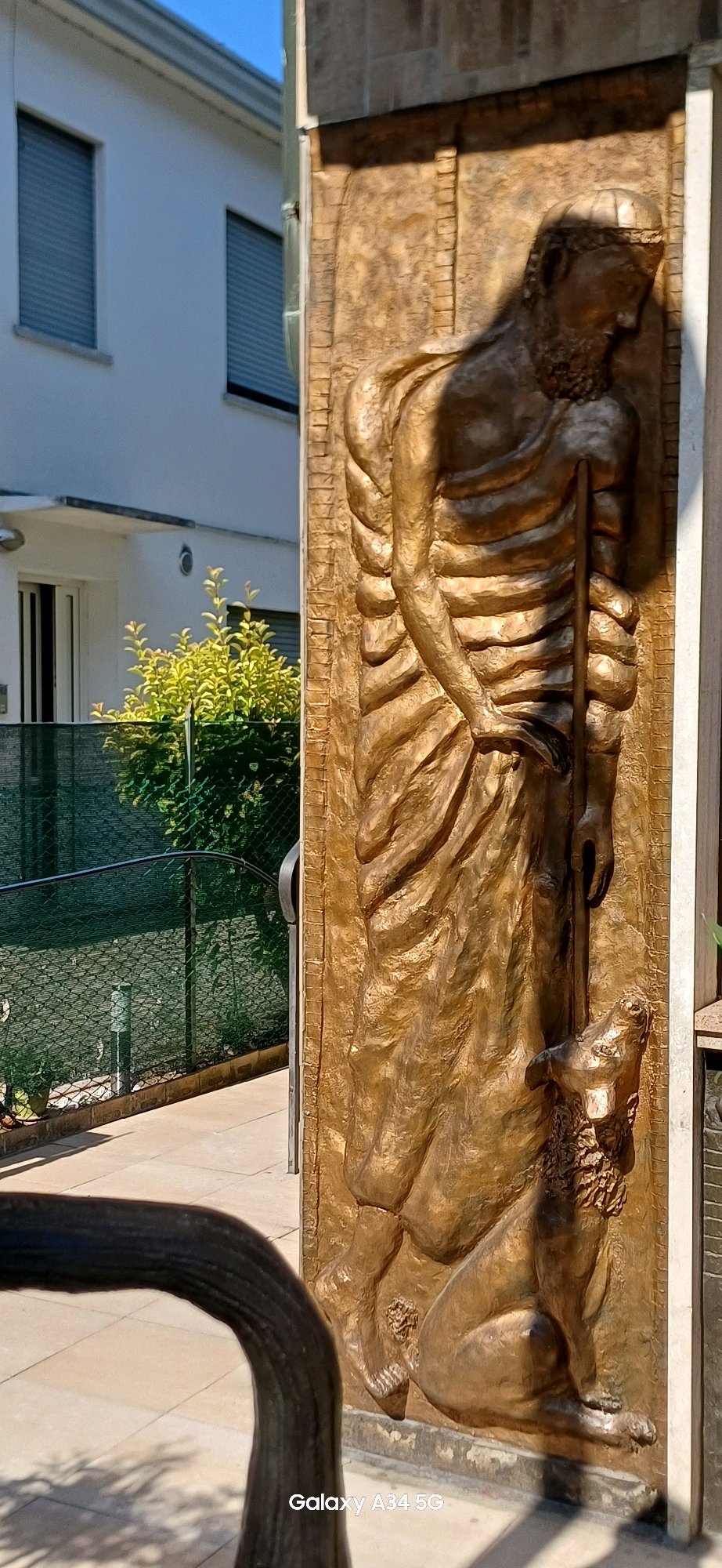 The image shows a relief sculpture depicting a man holding a staff and a dog. The sculpture is made of bronze and has a golden patina. The man is wearing a robe and has a beard. The dog is sitting at his feet and looking up at him. The sculpture is mounted on a wall, and it appears to be outdoors. In the background, there is a building with windows and a fence with green foliage. The scene is illuminated by sunlight, creating strong shadows. The lower right corner of the image shows a watermark that reads "Galaxy A34 5G". This sculpture is located in Abano Terme, Italy, specifically in the Piazza Cordellina, where it is part of a larger monument dedicated to the shepherd San Pancrazio.