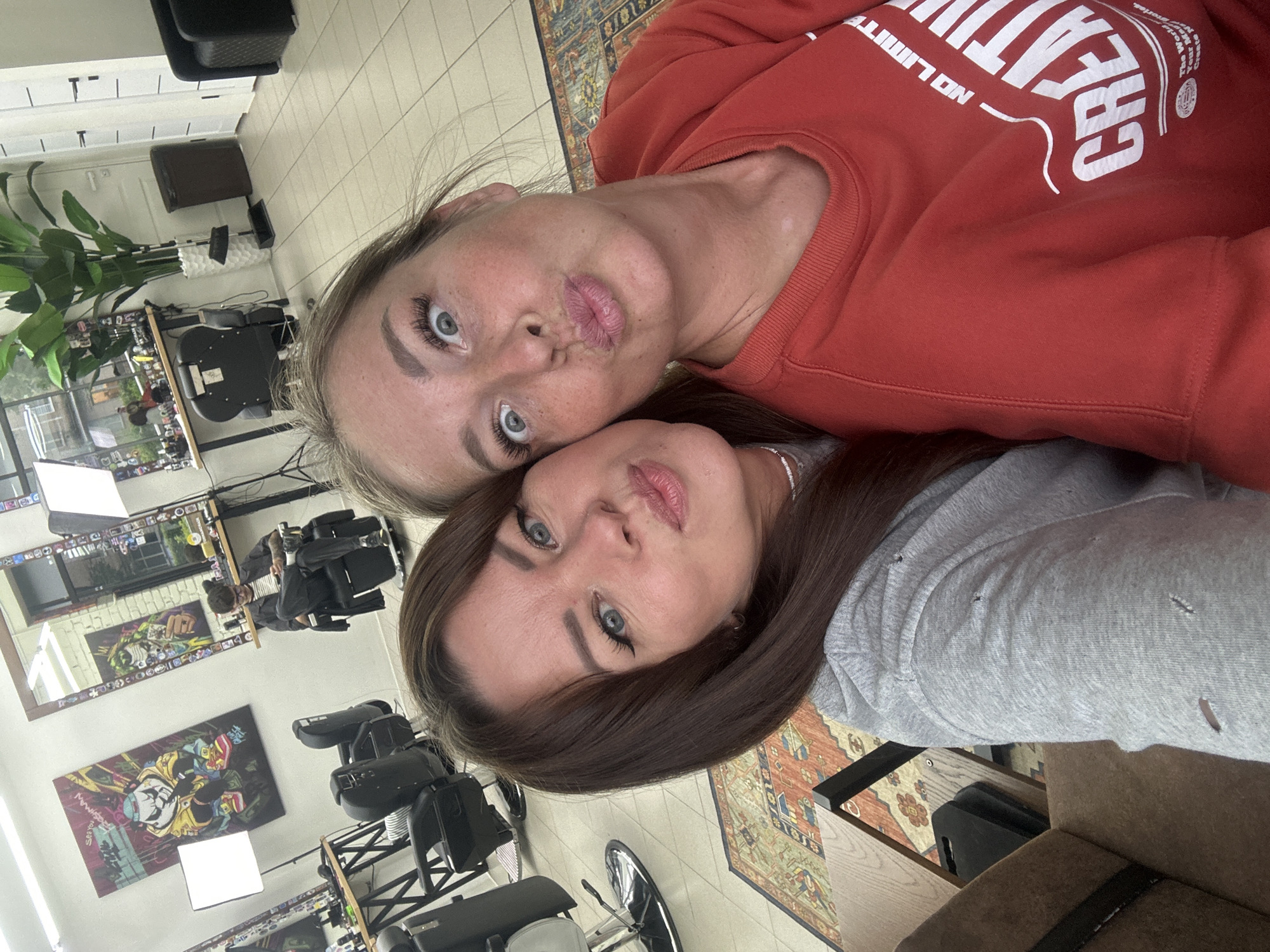 Two women are taking a selfie in a brightly lit barbershop. The woman on the left has long, dark brown hair and is wearing a gray hooded sweatshirt. She is looking at the camera with a pouty expression. The woman on the right has blonde hair and is wearing a red sweatshirt. She has her eyes wide open and is also making a pouty face. The background of the image shows several barber chairs, mirrors, and artwork on the walls. There is also a large potted plant and a trash can visible. The floor is tiled. The scene appears to be in a modern and stylish barbershop. The location is stated to be in Chaikovsky, Russia.