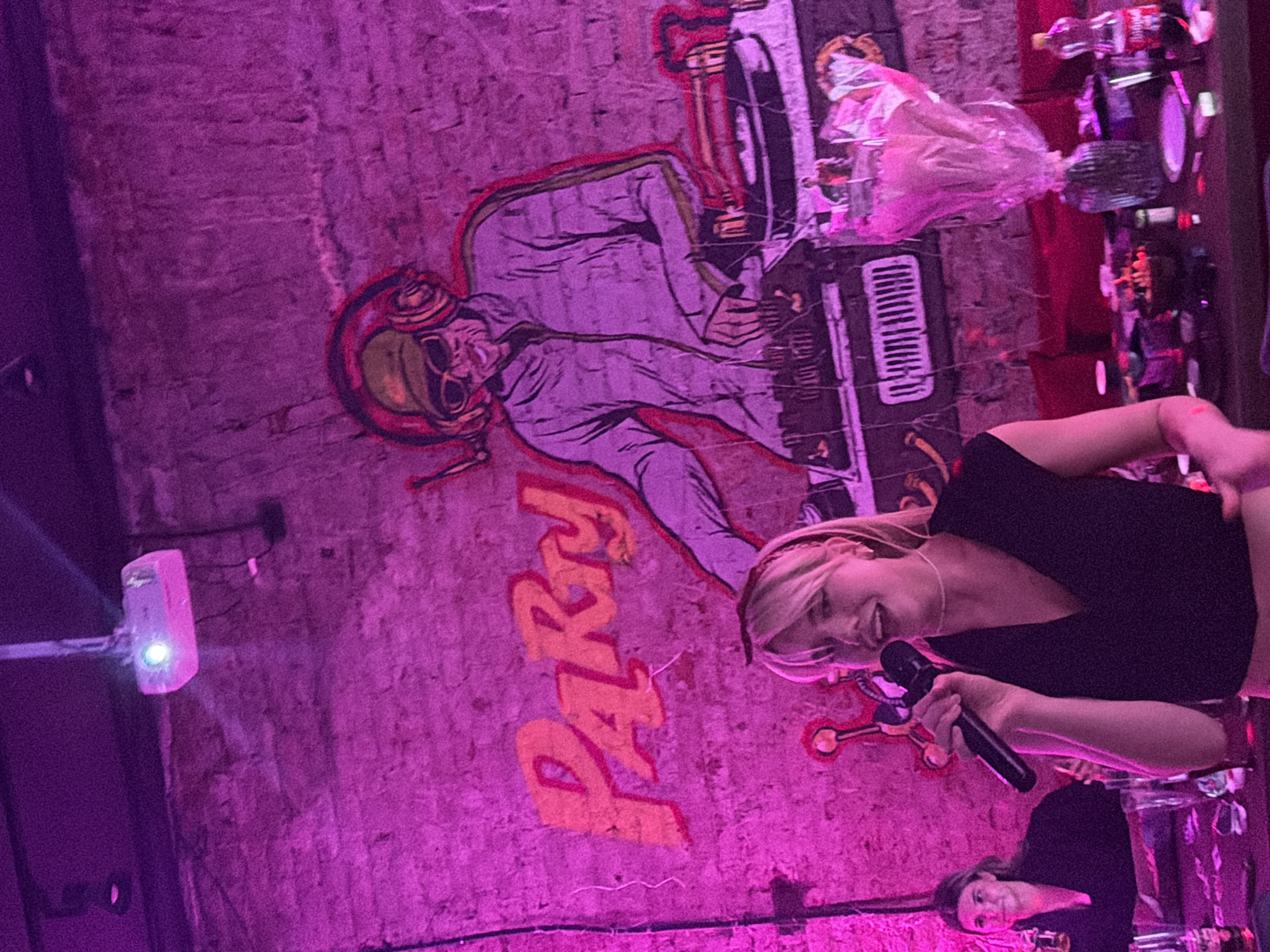 This is a candid, eye-level, medium shot photograph taken indoors under purple lighting. In the foreground, a young woman with blonde hair and bangs is singing into a microphone. She is wearing a black V-neck top and a thin white necklace. To her left, another woman with dark hair is sitting at a table, holding a drink. The background features a large mural on a brick wall depicting a DJ with headphones, sunglasses, and a record player, with the word "Party" painted in orange letters above it. A projector is visible on the ceiling. The table in the foreground is cluttered with drinks, snacks, and other items. The overall atmosphere appears to be a lively party or event.