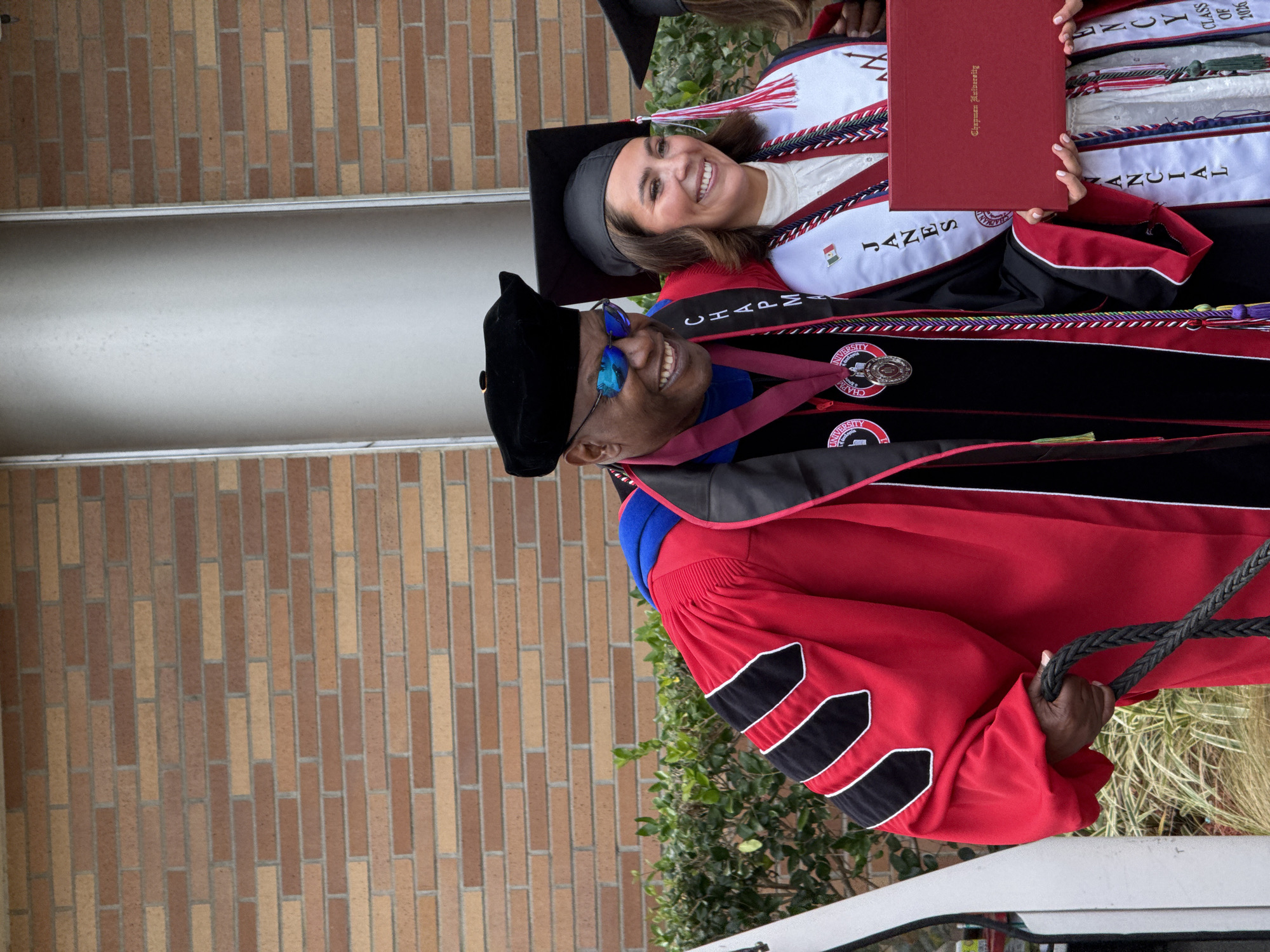 Here is a detailed description of the image:
**Scene**: An outdoor scene at Chapman University in Orange, California. The location is discernible by the presence of Chapman University regalia.
**People**: Two individuals are visible. One is a Black male wearing a black academic cap with a soft crown, sunglasses, and elaborate academic regalia, including a red robe and black stole. The other person is a fair-skinned female, wearing a graduation gown and holding a red diploma.
**Objects**: The primary objects are the graduation regalia (caps, gowns, stoles), a red diploma case labeled "Chapman University," a black braided cord held by the male figure, and what appears to be part of a vehicle in the foreground. The background includes a brick wall and greenery.
**Actions/Interactions**: Both individuals are smiling and appear to be posing for a photo. The man is looking directly at the camera while the woman is holding the diploma.
**Environment**: The background consists of a brick building and shrubbery, suggesting the photo was taken on the university campus. The lighting seems to be bright and outdoors.