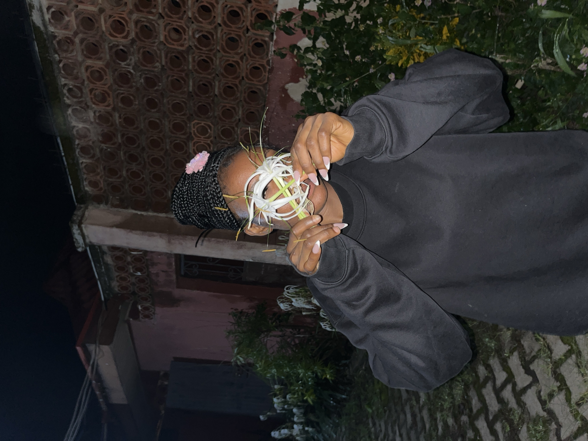 A young Black woman stands outdoors at night, holding a white flower with long, thin petals over her eyes. She is wearing a black crewneck sweatshirt. Her hair is braided and adorned with a small pink flower. Behind her, a textured brick wall with circular openings is visible, with a portion of a pink building to the left. The ground is paved with interlocking bricks. In the background, there is lush green foliage with some white and pink flowers. The scene is illuminated by artificial light, creating a dark and moody atmosphere. This appears to be a residential area in Jos, Nigeria, possibly in a courtyard or garden.