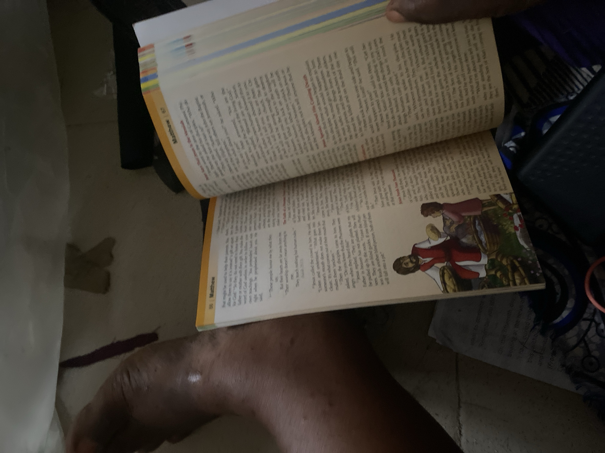 The image shows a person's leg and foot positioned next to an open book. The book appears to be a Bible, with text and illustrations visible. The person's leg is dark-skinned and appears to be resting on or near a surface with a patterned fabric. The background is dimly lit and out of focus. The context of Jauro Yinu, Nigeria, is not present in the visual information of the image.