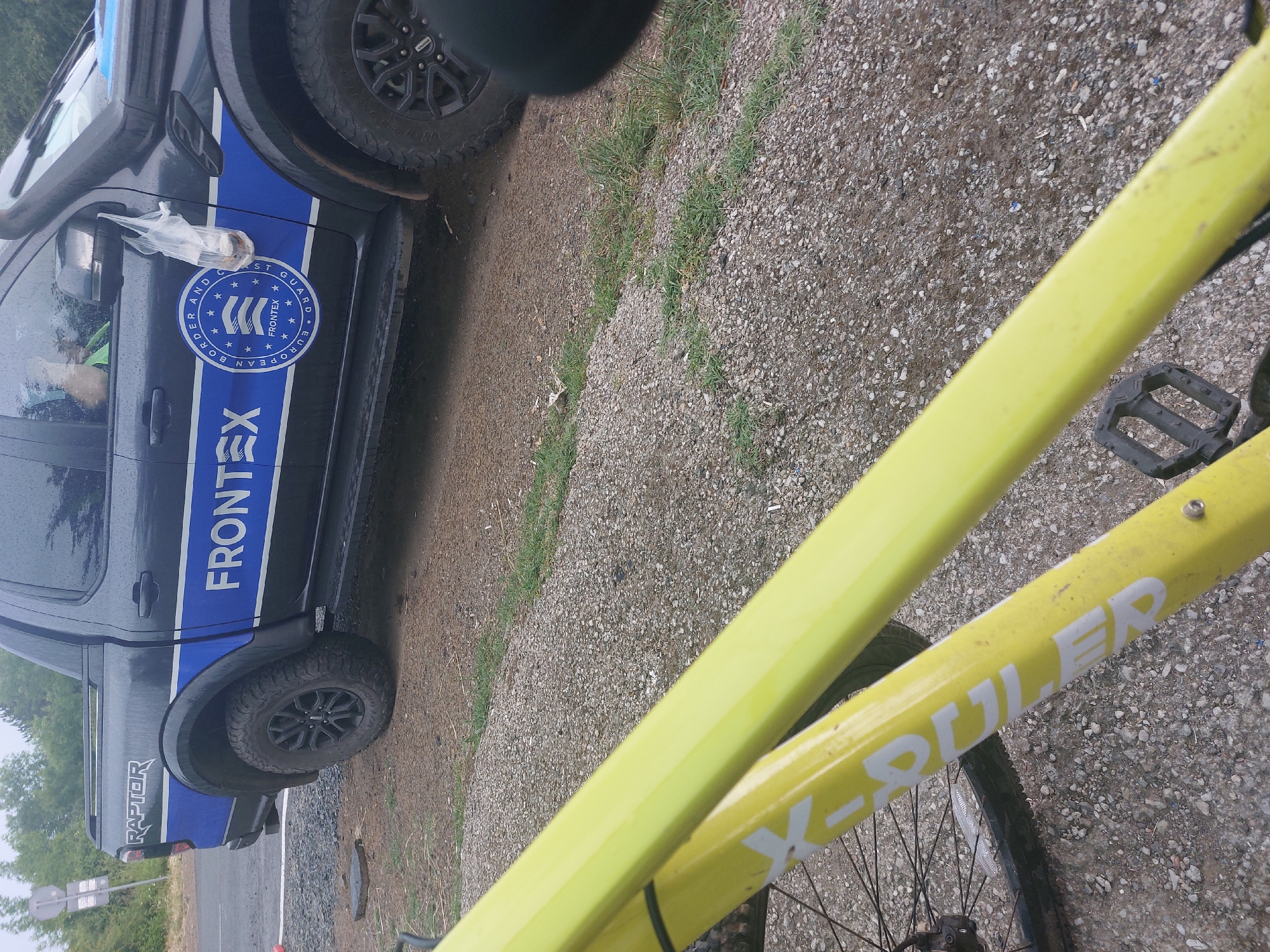 The image shows a FRONTEX vehicle parked on what appears to be a gravel road or shoulder, possibly in a rural or semi-rural area. A yellow bicycle partially obscures the foreground. The FRONTEX vehicle is a dark-colored pickup truck with the agency's logo and branding clearly visible. The road surface is light-colored gravel, with some grass growing along the edge. Based on the context, it can be inferred that the image may be connected to FRONTEX's activities related to border control or monitoring in the area. There is no indication of Tsarevo, Bulgaria, directly in the image, so it cannot be concluded that it is the location.