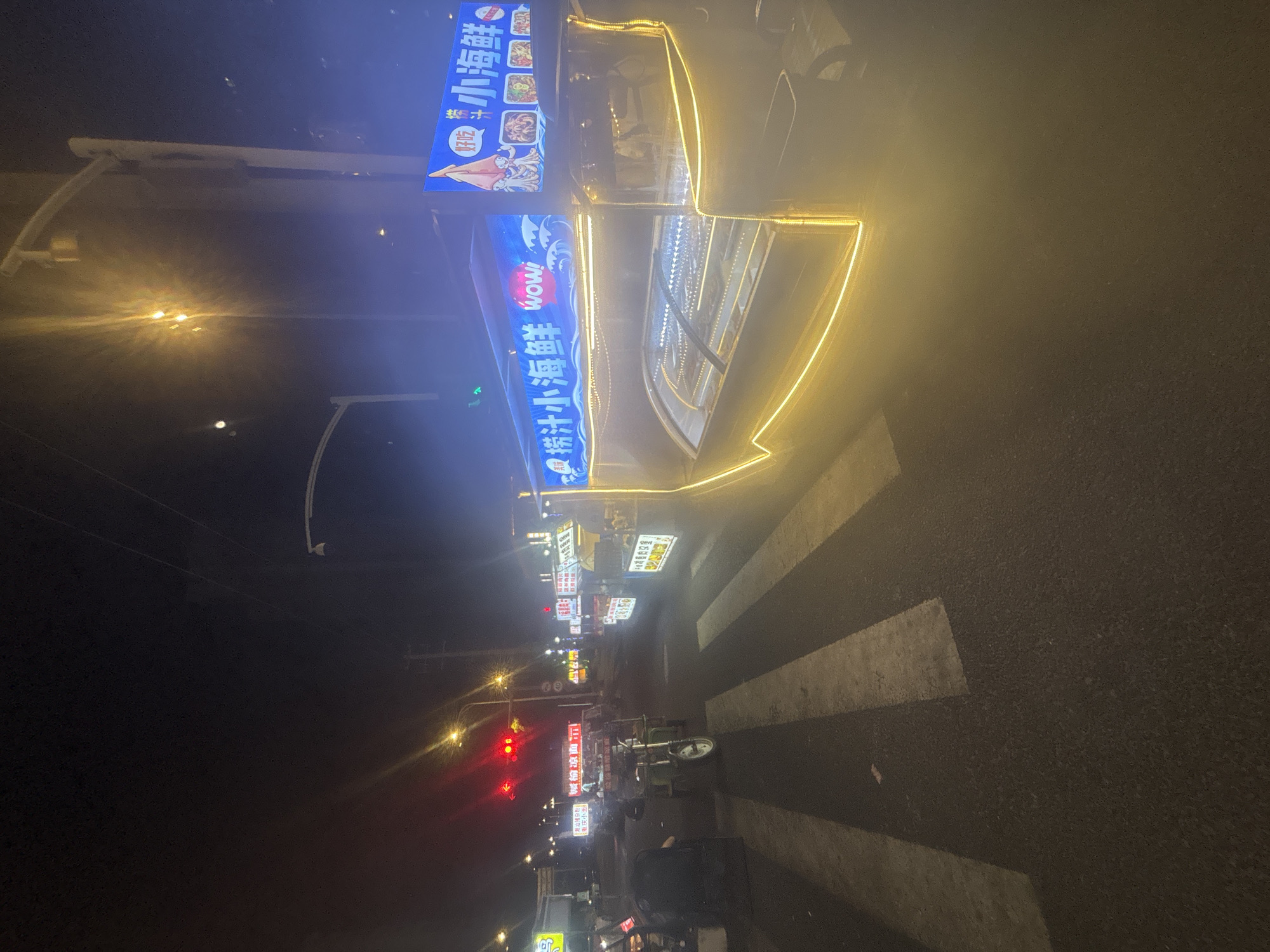 The image depicts a night scene on a street in Suzhou Industrial Park, China, likely a commercial area with food vendors. A prominent food stall with bright blue and yellow neon lighting is the central subject. The stall has a sign that reads "捞汁小海鲜" (Luō zhī xiǎo hǎixiān), which translates to "Salty Juice Seafood." The stall features a display case with what appears to be seafood, illuminated from within. The stall is decorated with a yellow neon outline along its edges.
In the foreground, a pedestrian crossing is visible, with white stripes on the dark asphalt road. To the left of the food stall, other stalls and vehicles are present, though less distinct due to the lighting and distance. A traffic light showing red is visible further down the street, indicating traffic control. The overall atmosphere is one of a bustling night market or street food scene. The lighting conditions suggest it is nighttime, with artificial lights from the stalls and streetlights illuminating the area. There are no people clearly visible in the image.