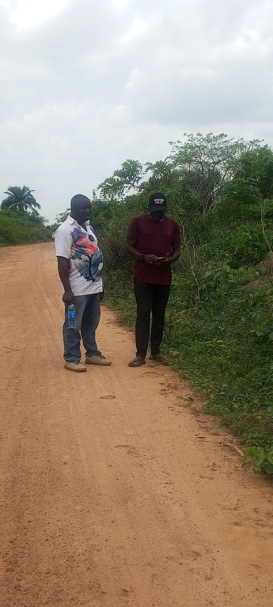 The image depicts two individuals standing on a dirt road in Otunade, Nigeria. The man on the left is wearing a white shirt with a graphic design, blue jeans, and brown shoes. He is holding a bottle of water. The man on the right is wearing a maroon shirt, black pants, and a black baseball cap with "GOOD 2027" printed on it. He is looking at a handheld device, possibly a phone. The road is lined with green vegetation on the right side. The sky is overcast.