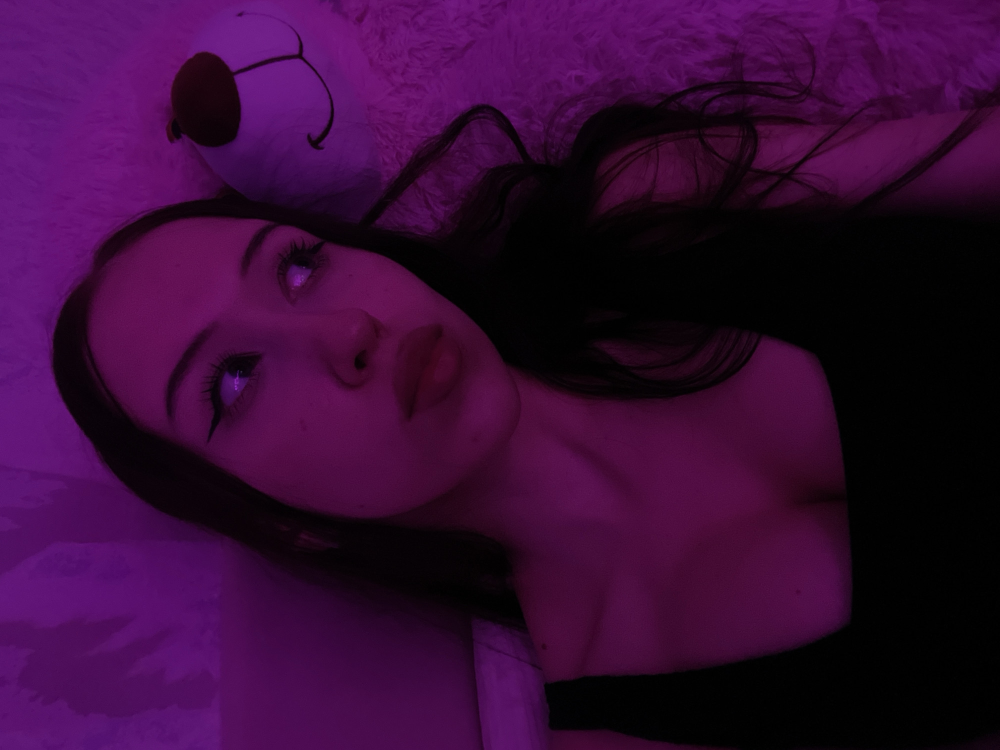 The image shows a young woman with long dark hair lying down, illuminated by purple light. She is wearing a black top. To her right is a white teddy bear with a red nose and embroidered eyes and mouth. The background appears to be a textured surface. There is no information to indicate the location is Votkinsk, Russia.