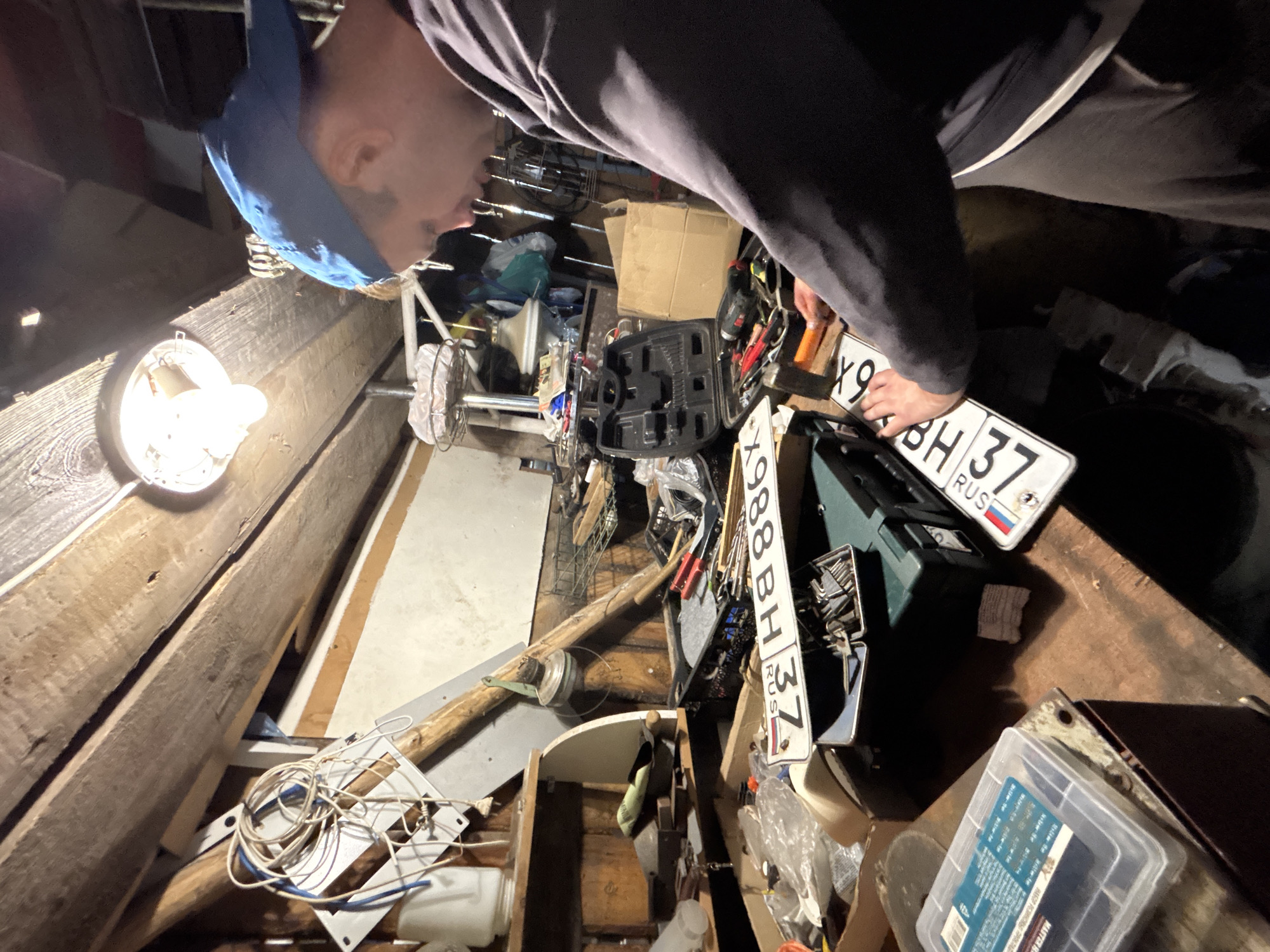 A man wearing a blue baseball cap and dark long-sleeved shirt works in a cluttered workshop. He is standing at a workbench, holding what appear to be Russian license plates. The workshop is dimly lit by an overhead lamp and filled with various tools, boxes, and miscellaneous items. The walls and ceiling are made of wood, suggesting a rustic or older structure. The overall impression is one of a working space dedicated to manual tasks, likely in a rural or semi-rural setting such as Dunilovo, Russia.