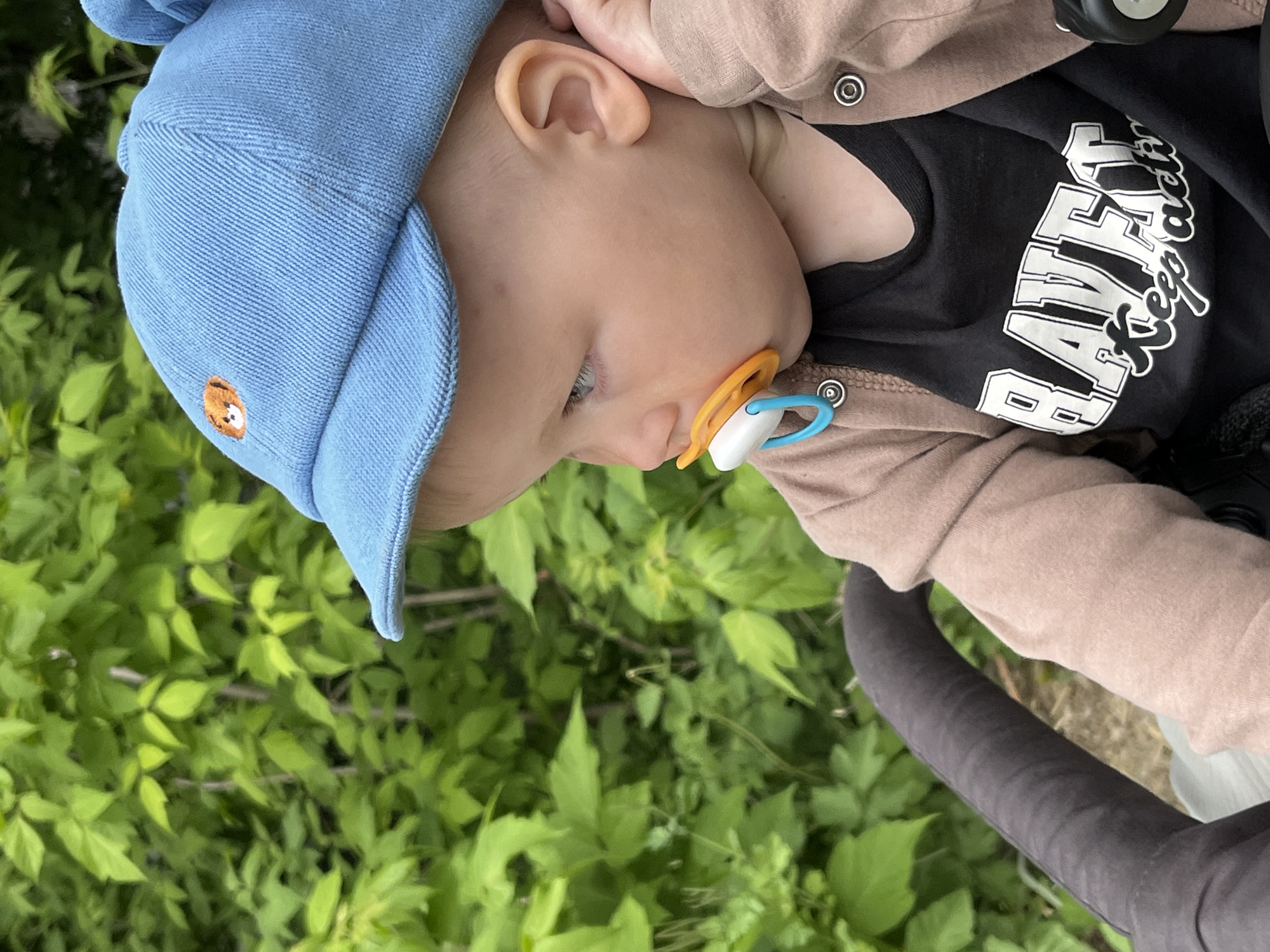 A baby wearing a blue hat and holding a pacifier is lying in a stroller. The baby is wearing a black shirt with white lettering and a beige jacket. The stroller is on a paved surface near green vegetation. The exact location in the city of Chaikovsky, Russia is indeterminable.