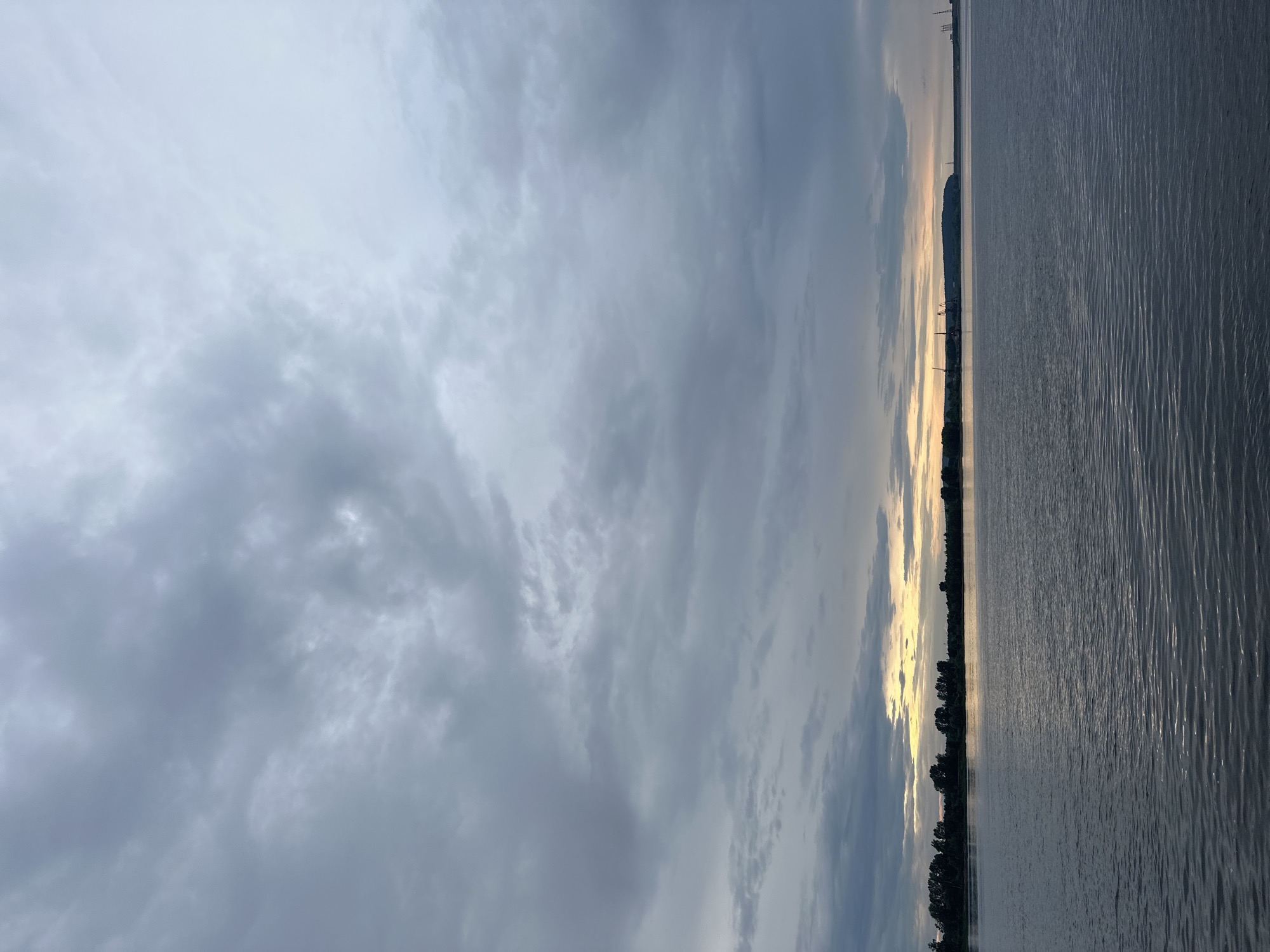 The image depicts a wide, calm body of water, likely the Kama River, as viewed from the embankment in Chaikovsky, Russia. The sky is overcast with heavy, grey clouds, but a sliver of yellowish light breaks through at the horizon, suggesting either dawn or dusk. The distant shoreline is lined with a dense band of dark green trees, and beyond them, in the right portion of the frame, industrial structures including cranes are visible, hinting at a port or industrial area. The water's surface is gently rippled, reflecting the muted light of the sky. There are no people or animals visible in the scene. The overall mood is somber and tranquil, with a touch of industrial presence on the horizon.