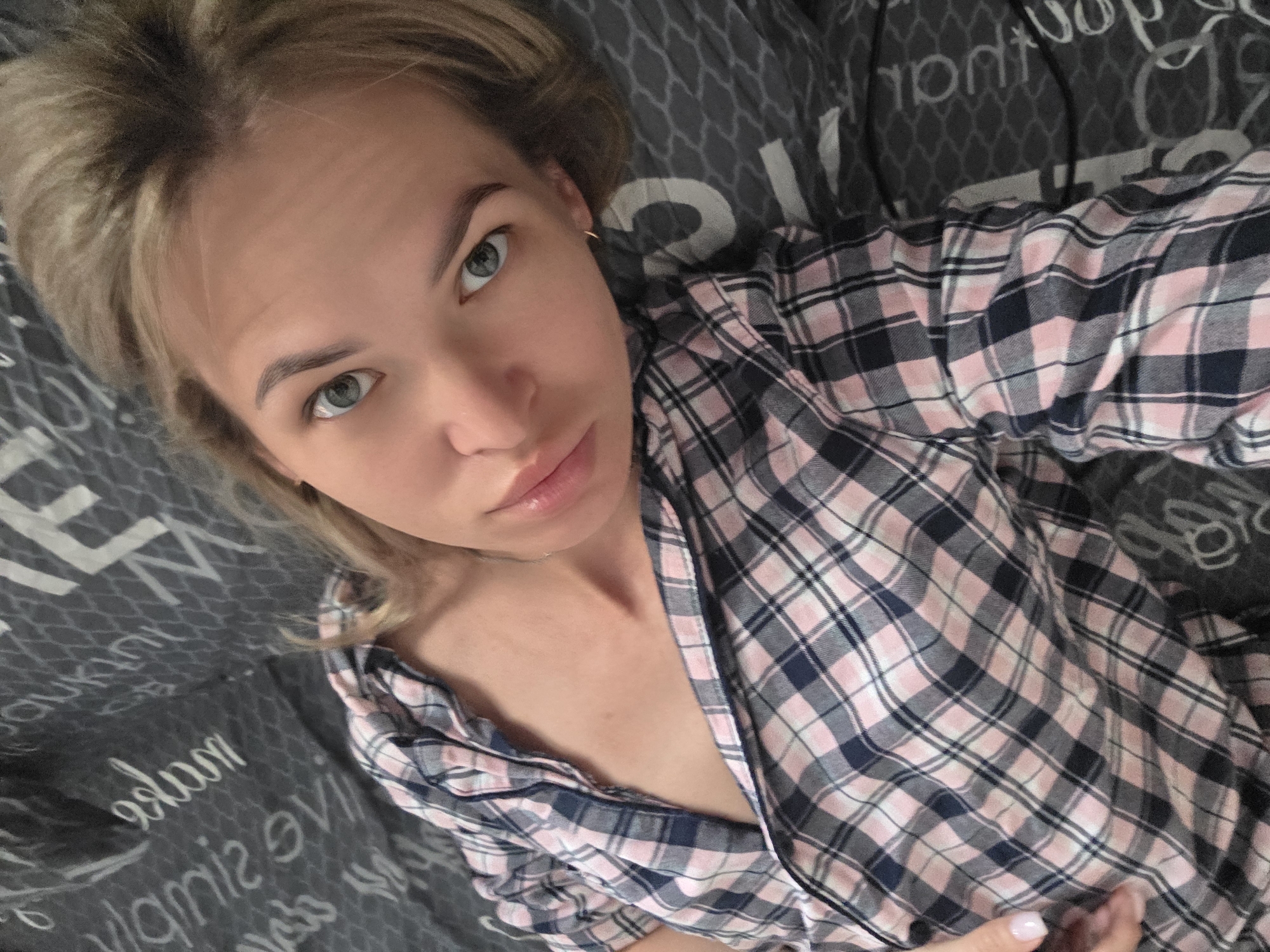 A young adult female with blonde hair and blue eyes lies on a gray patterned bed or sofa. She is wearing a pink, white, and navy blue plaid shirt, which is unbuttoned at the top. Her skin is fair, and she appears to have a small gold hoop earring. The background consists of the gray bedding with what appears to be repeating white text. The image appears to be a selfie taken at a close angle. There is no information available in the image about the location being in the city of Chaikovsky, Russia.