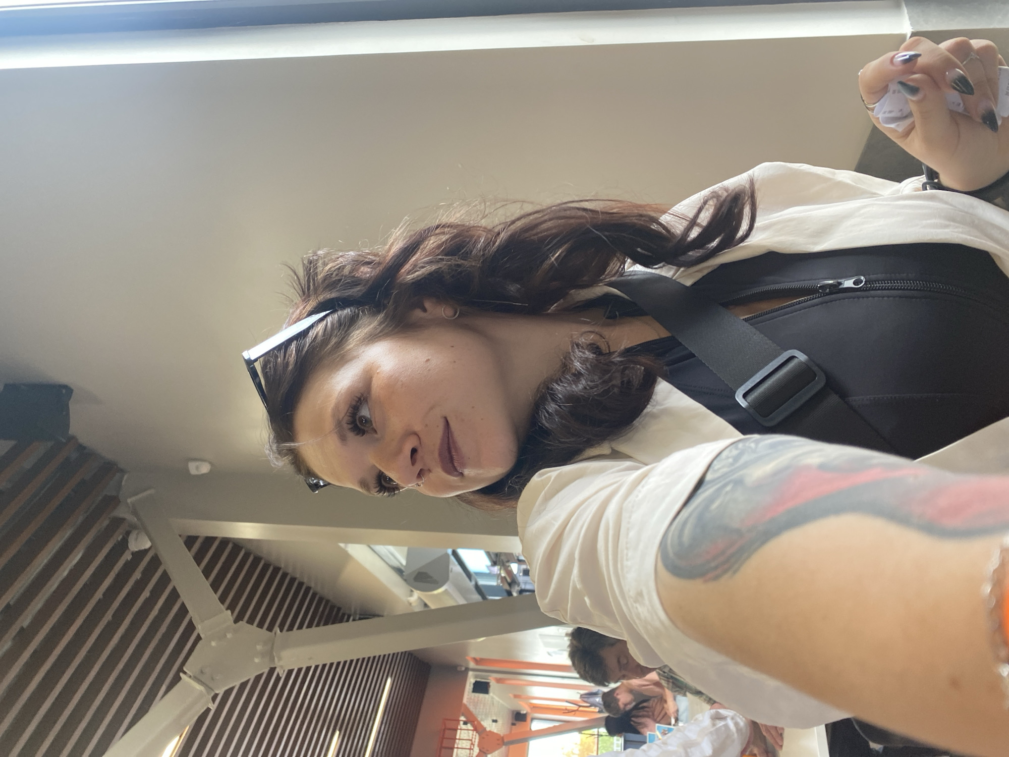 In an indoor setting that appears to be a cafe or restaurant, a woman with long, dark wavy hair, wearing a white open jacket over a black zippered top and sunglasses perched on her head, is looking upwards and to her left. She has prominent eyelashes and is holding a crumpled piece of paper in her right hand, which has dark ombre fingernails. A black strap from a bag is visible across her chest. Her left arm, which is tattooed, is in the foreground. In the background, other patrons are seated at tables, with architectural elements including wooden slats on the ceiling and white structural beams visible. The scene appears to be consistent with an interior establishment in Votkinsk, Russia.