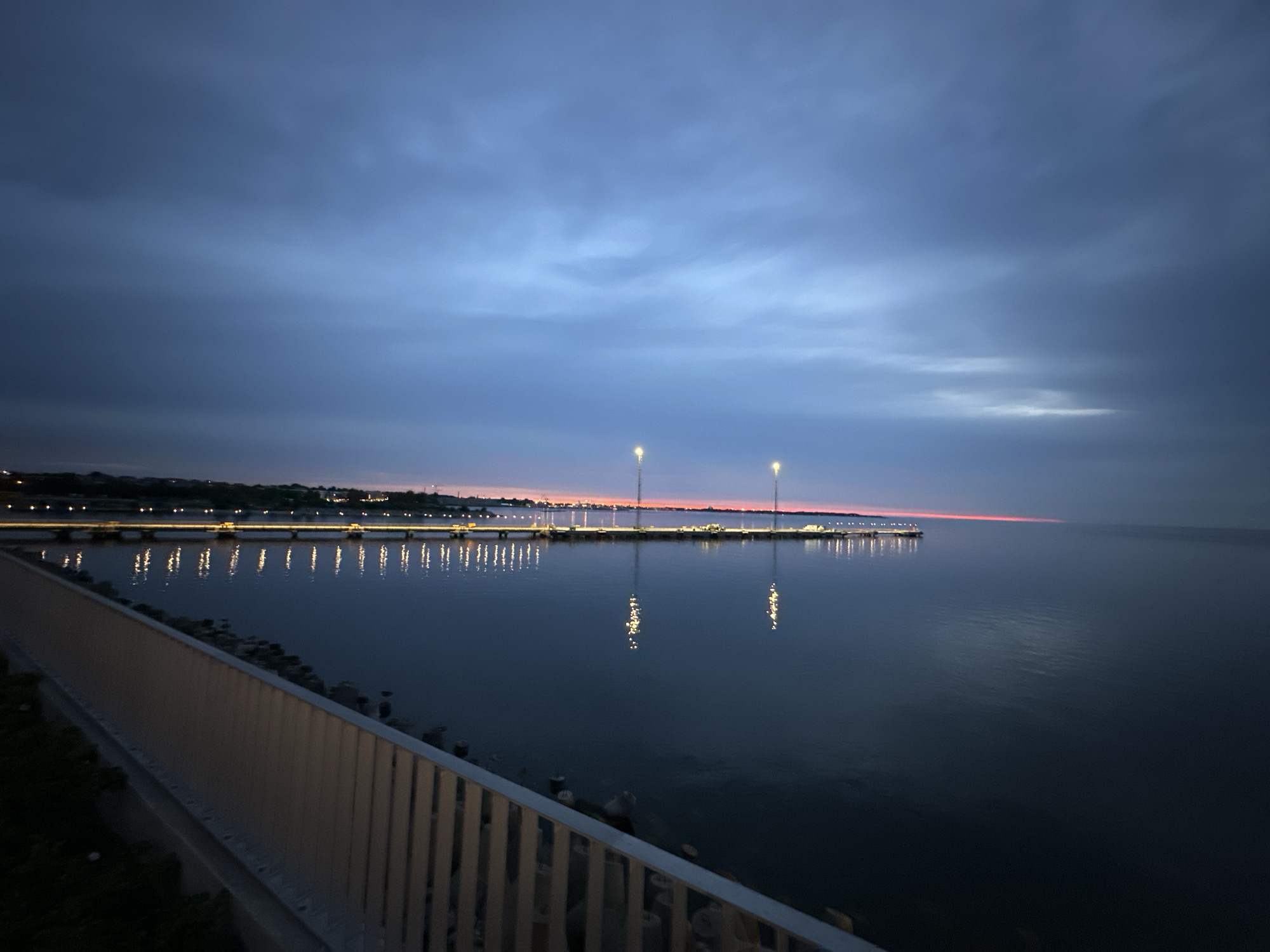 The image depicts a coastal scene at dusk, likely in Tallinn, Estonia, given the distinct architectural and harbor features. The foreground features a modern-looking metal railing and concrete pathway, suggesting a waterfront promenade or park. To the right, a vast expanse of dark, calm water stretches towards the horizon.
In the middle ground, a long pier or breakwater extends into the sea. It is illuminated by numerous lights, which cast reflections on the water's surface, creating a shimmering effect. Several tall light poles are also visible along the pier. Beyond the pier, a faint line of lights suggests buildings or a coastline, possibly the city of Tallinn. The sky is a gradient of deep blues and grays, with a thin band of pink and orange visible on the horizon, indicating the last vestiges of sunset or twilight. The overall atmosphere is serene and tranquil.