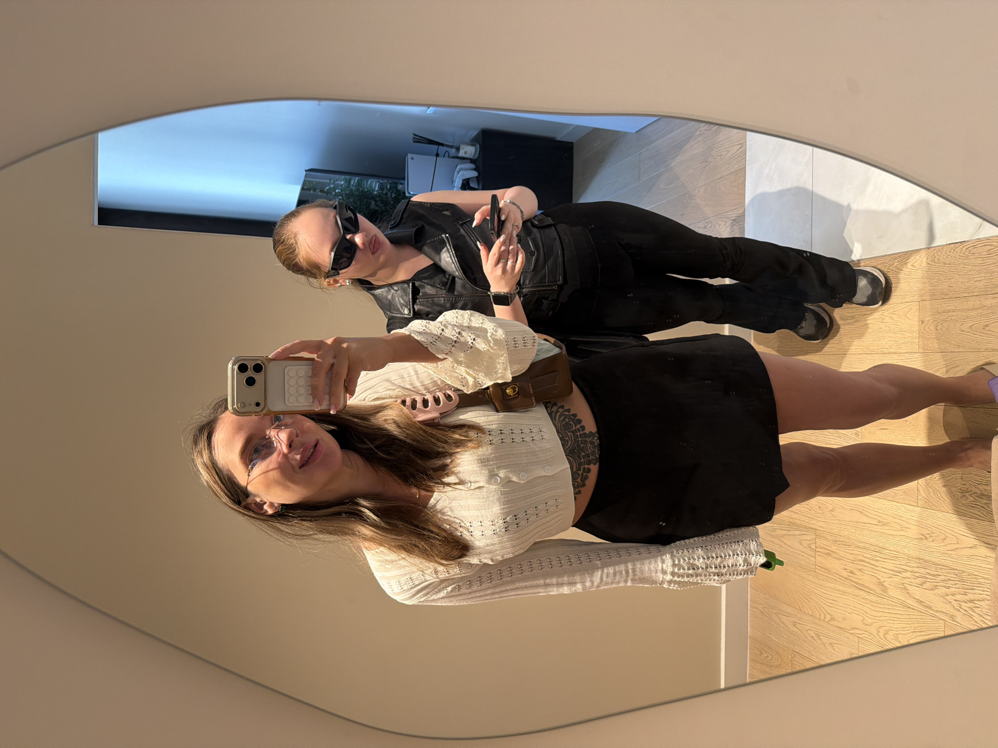 Two women are reflected in a large, oval mirror. The woman on the left is facing the mirror, holding a smartphone in her right hand, taking a selfie. She has long, blonde hair, is wearing glasses, a white crocheted cropped cardigan, and a black mini-skirt. She has a tattoo on her stomach. She is holding a small brown purse on her left shoulder and a green object in her left hand. The woman on the right is standing behind and to the right of the first woman, looking at her phone. She has blonde hair pulled back, is wearing large black sunglasses, a black sleeveless leather jacket, and black pants. The background of the reflection shows a room with wooden floors and some furniture, including a black cabinet with items on it. There is no indication of this scene being in Moscow, Russia.
