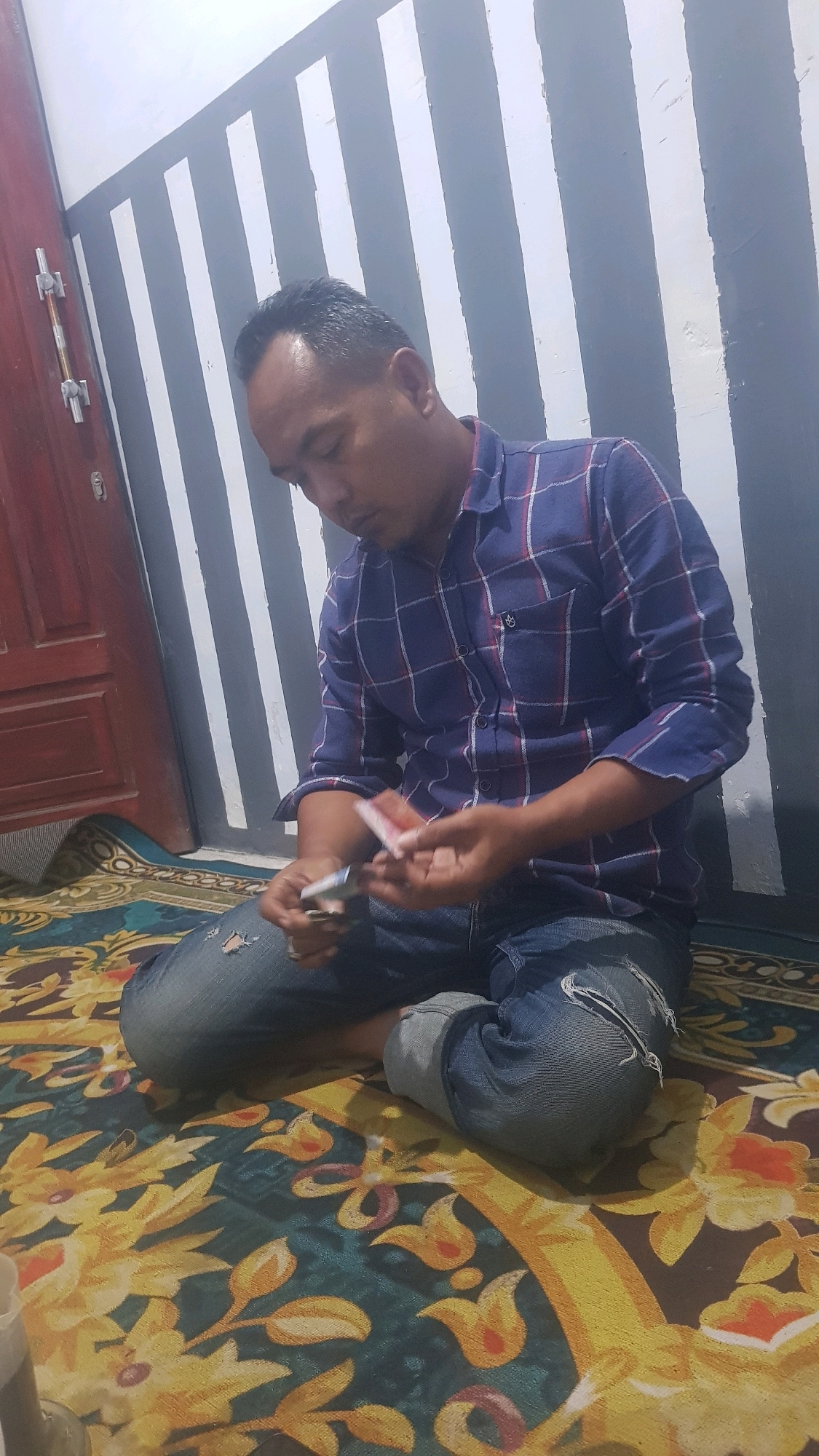 A man is sitting on a patterned carpet, with his legs crossed. He is wearing a blue and red plaid shirt and ripped blue jeans. He is holding what appear to be playing cards or similar items in his hands, and is looking down at them. In the background, a wall with vertical black and white stripes can be seen. To the left, a portion of a dark red door is visible. The scene appears to be indoors, likely in a residential setting in Bakalan, Indonesia.