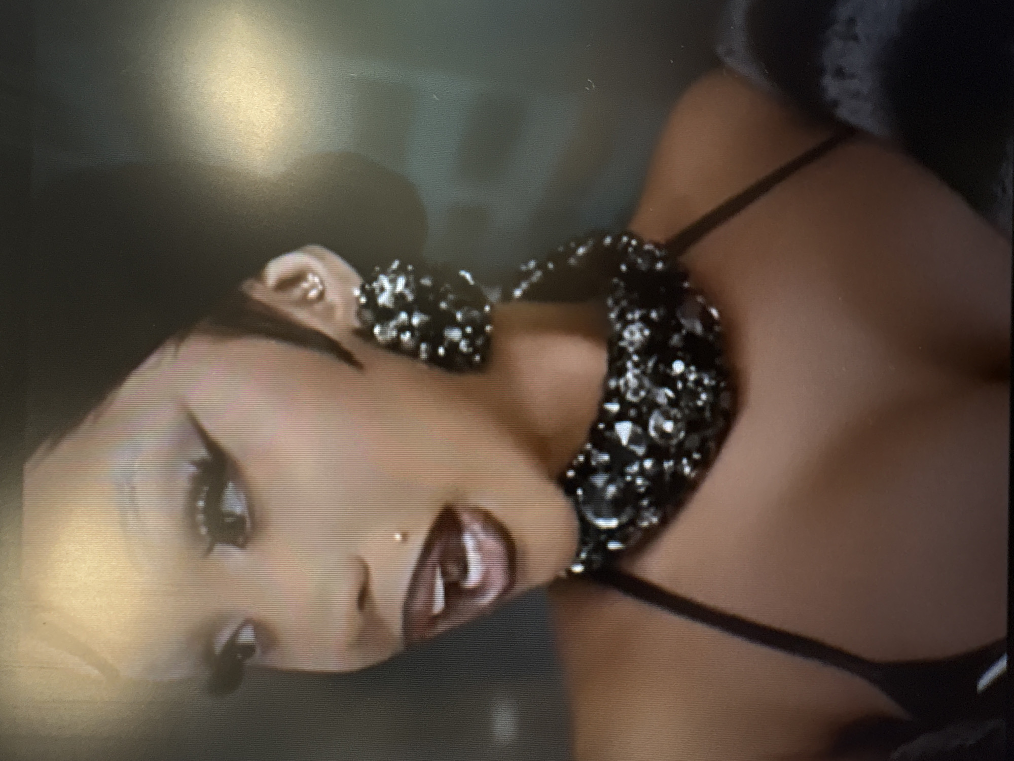 The image is a close-up shot of a person's face and upper body, with a focus on their makeup, jewelry, and clothing. The person has dramatic eye makeup, including sharp eyeliner and long eyelashes. They are wearing large, sparkling earrings and a statement necklace. Their lips are painted with a dark lipstick, and they have a small stud piercing on their philtrum. The person is wearing a black top with thin straps, and the background is dark and somewhat out of focus. There is no indication of the location being Sofia, Bulgaria; the scene appears to be indoors, possibly a studio or a dimly lit room.