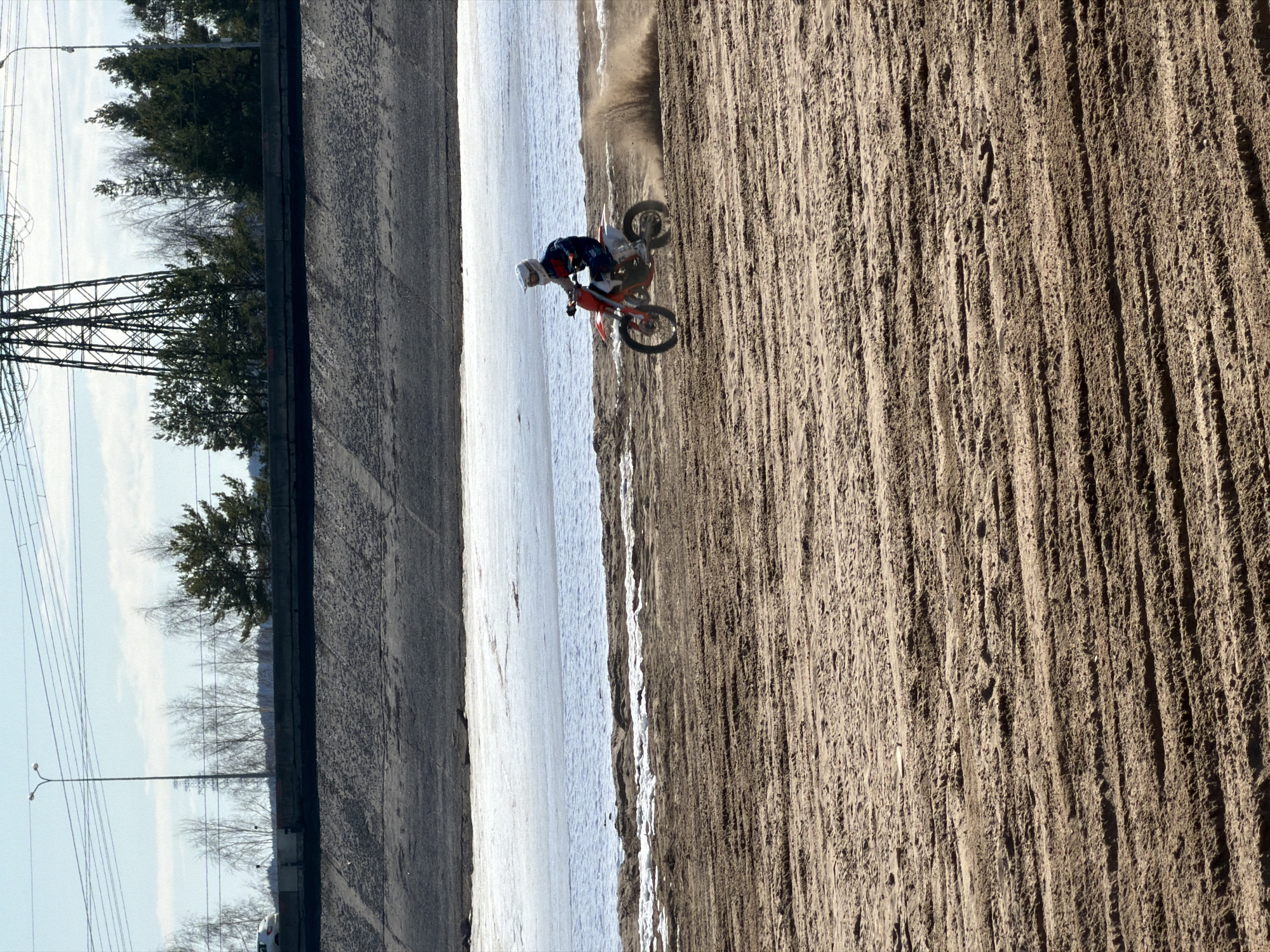 A motorcyclist rides on a dirt track. Trees, lampposts, and powerlines are visible in the background. The location is Noviy, Russia.
