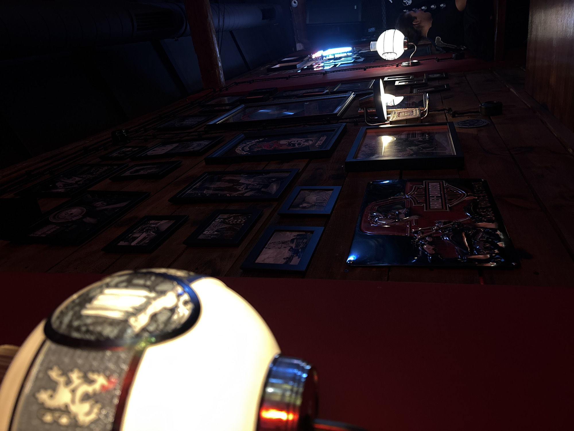The image shows an interior wall decorated with numerous framed photographs and Harley-Davidson memorabilia. Two lamps are visible, one in the foreground and another further back. The wall appears to be made of wood and painted red. The photographs are in dark frames. The lighting is dim. The specific location within Belgrade, Serbia cannot be determined from this image alone.