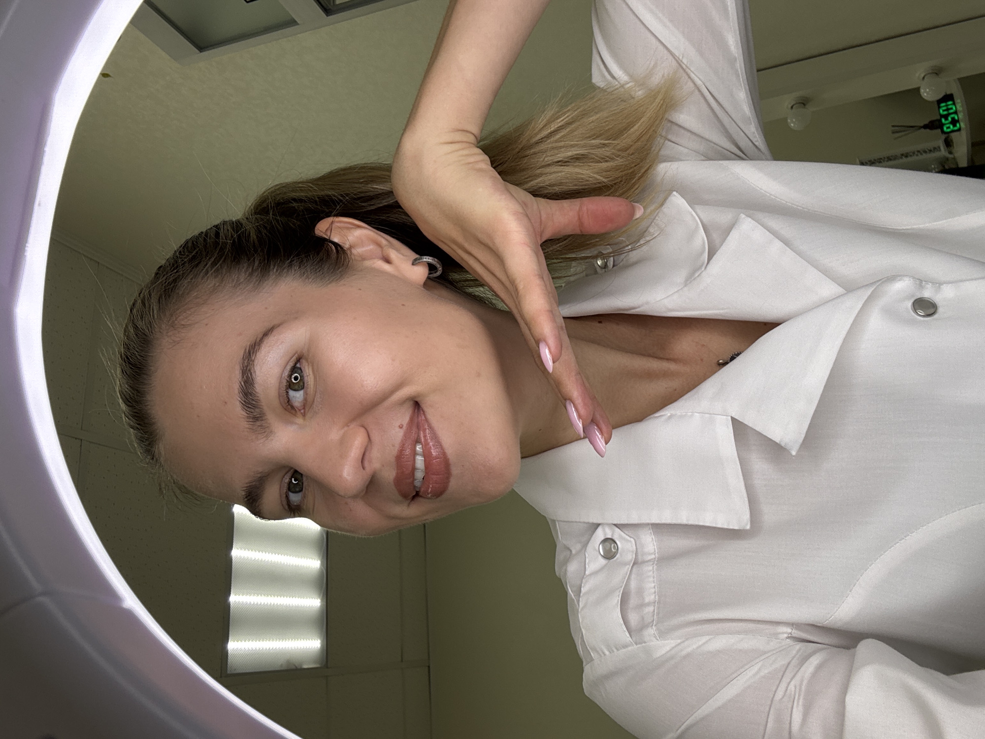 A blonde woman with fair skin, green eyes, and her hair in a ponytail is looking at the camera and smiling. She is wearing a white button-up shirt. Her right hand is raised to her face, with her fingers resting on her chin and cheek. The background appears to be an indoor setting with a fluorescent light fixture on the ceiling and a ring light illuminating the woman. A digital clock displaying "10:51" is visible in the background, along with a diffuser and some light bulbs.