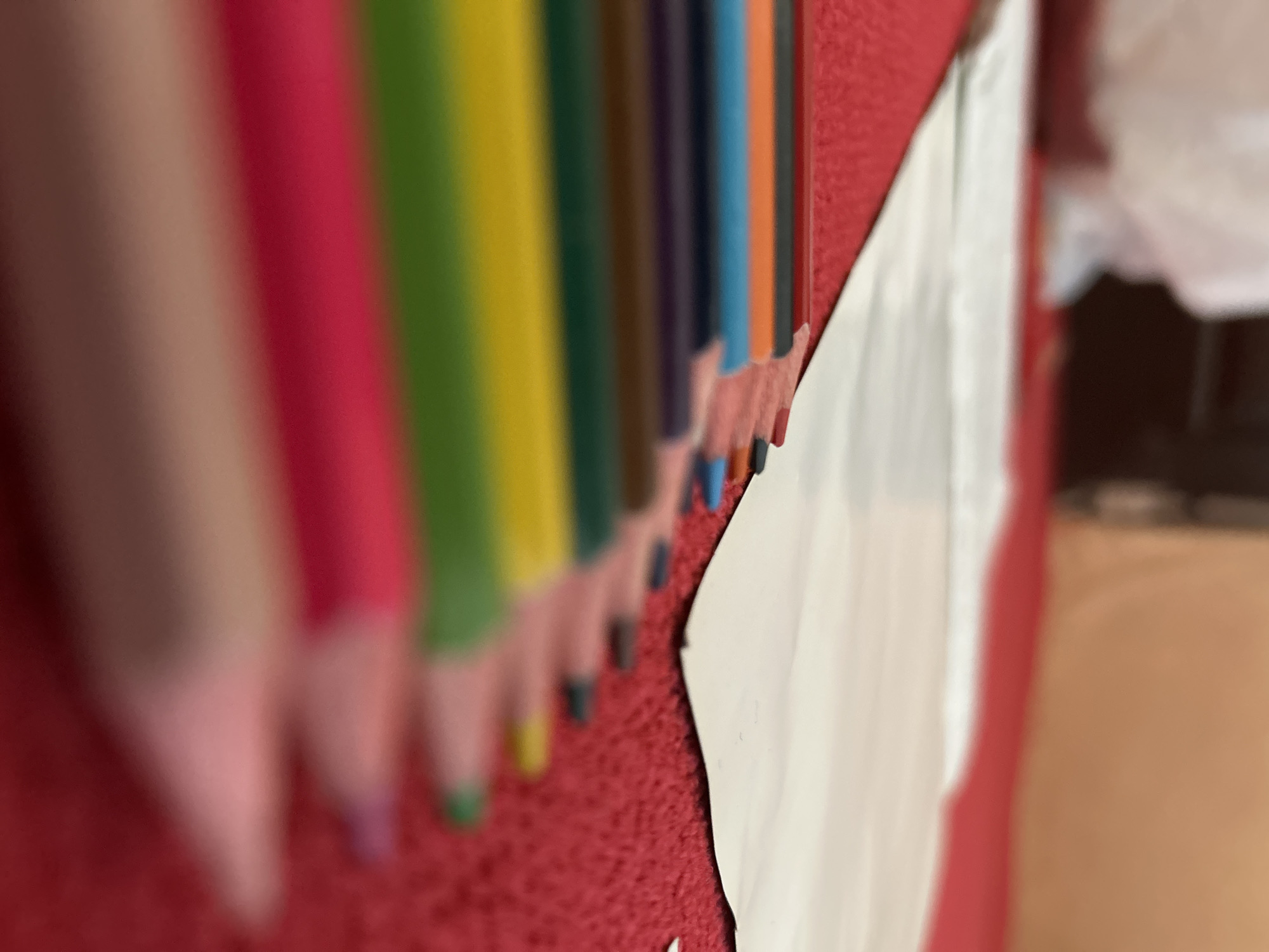 The media file displays an abstract, close-up, and angled composition of an array of colored pencils positioned against a vibrant red, textured surface. The pencils are arranged in a row, appearing diagonally across the frame due to the shot angle, showcasing various colors including pink, red, green, yellow, brown, purple, blue, and orange. The sharpened tips of several pencils are visible, resting on or near the red surface. The pencils on the far left are significantly blurred, while those towards the center and right maintain more focus.
To the right of the pencils, a piece of white paper with an uneven, possibly torn or cut, edge is affixed to the same red, textured background. This red surface appears to be a consistent, finely textured material, such as carpet or felt. The background beyond the pencils and paper is heavily out of focus, revealing indistinct light and dark areas consistent with an indoor environment.
No people are visible, and there is no specific activity or event being depicted beyond the static arrangement of the objects. The image is a still life focusing on the colors and textures. There is no visible text. The scene is set indoors in Lhokseumawe, Indonesia, characterized by its warm color palette and shallow depth of field.