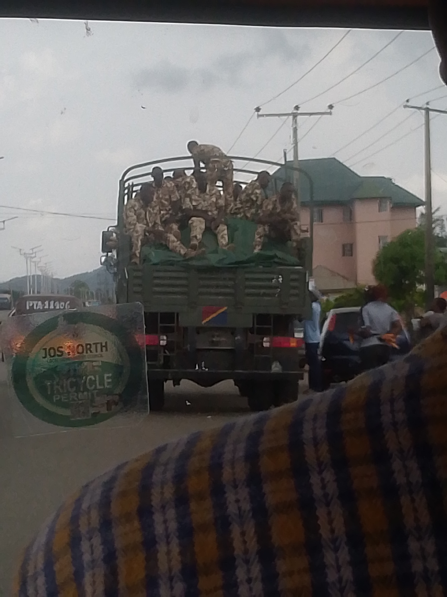 The image shows a military truck in Bukuru, Nigeria, carrying numerous soldiers. The truck is green and has a Nigerian flag displayed. Soldiers are visible in camouflage uniforms, some standing and some sitting on top of the truck. The truck appears to be stopped in traffic, with civilians and other vehicles nearby. A building with a green roof is also visible in the background. A "Jos North Tricycle Permit" sticker is affixed to the window through which the photo was taken. Another vehicle in the image has a license plate that reads "PTA-11406."
