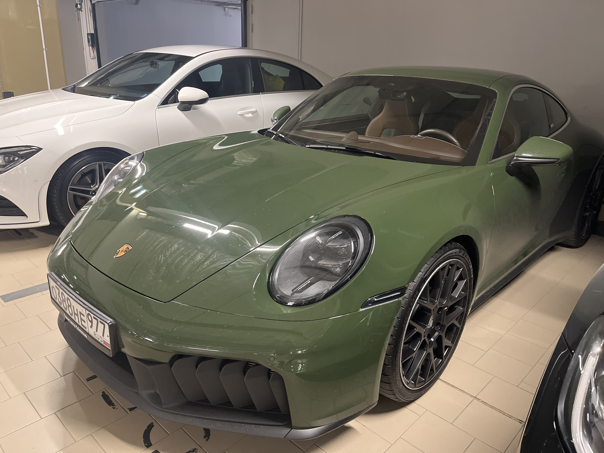 The image shows two cars parked indoors, likely in a garage or a showroom. In the foreground, a dark green Porsche 911 is parked, facing slightly to the left. The car has a sleek design with prominent headlights and a sporty bumper. Its interior appears to be light brown or tan. A license plate is visible on the front bumper, displaying the text "0388HE977 RUS".
Behind the Porsche, a white sedan is parked, also facing left. Its front end is partially visible. To the right of the Porsche, the front of a black car is visible, its headlights illuminated. The flooring of the scene consists of light-colored tiles. The lighting suggests it is indoors, and there is no indication of the time of day or weather conditions. The overall scene conveys a sense of automotive display or storage.