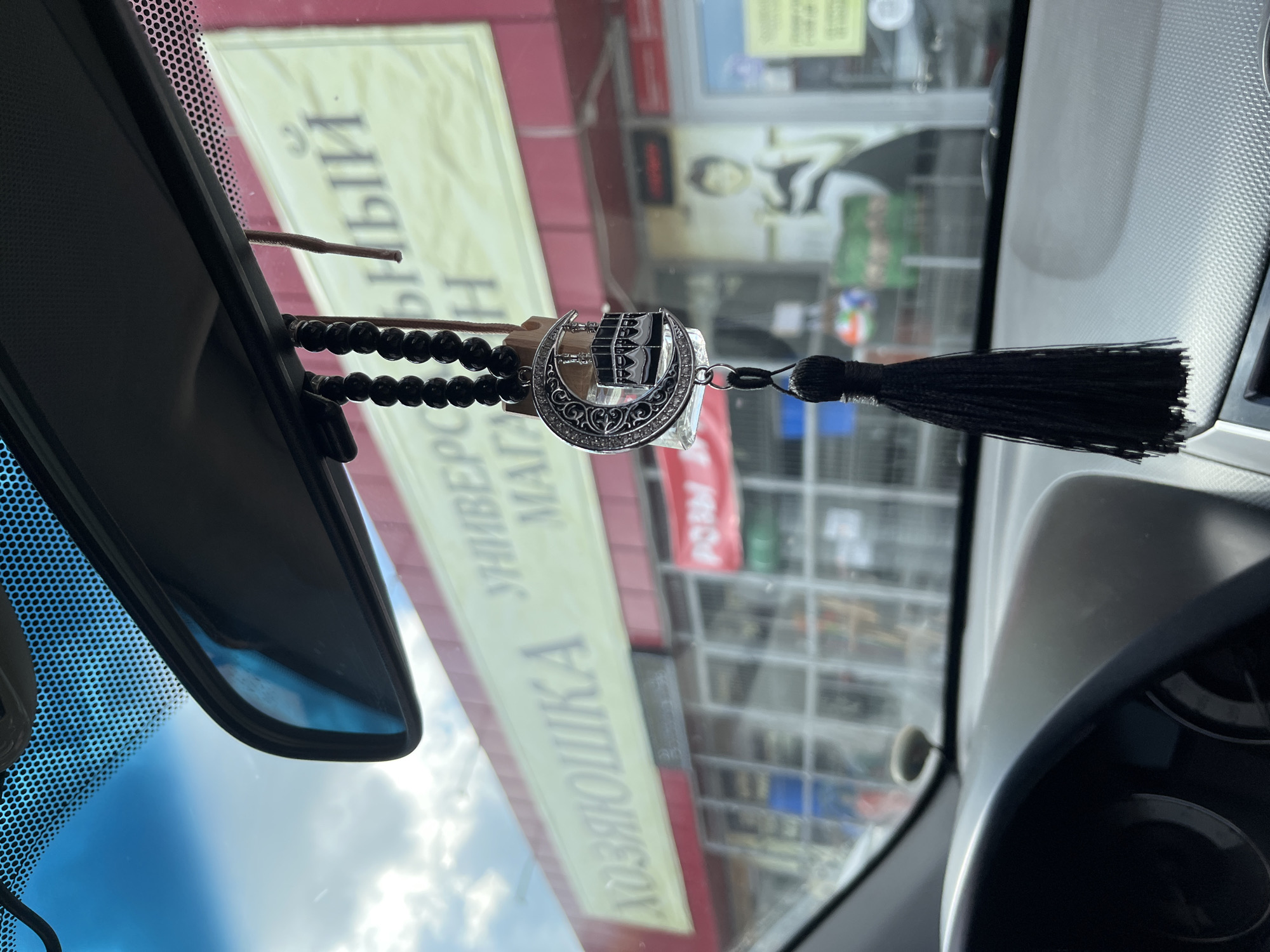 The image was taken from inside a vehicle, looking out through the windshield. In the foreground, a black tassel hangs from a string, attached to a keychain that features a crescent moon with an Islamic symbol of the Kaaba. Behind the keychain, out of focus, is a blurred yellow sign on a building that reads in Russian "ХОЗЯЮШКА" (Khozyayushka) and "УНИВЕРСАЛЬНЫЙ МАГАЗИН" (Universal Store). The building has large windows and a red awning. The sky is visible above the sign with some clouds. The dashboard of the car is visible in the lower part of the image. The location is specified as Novaya Otradovka, Russia.