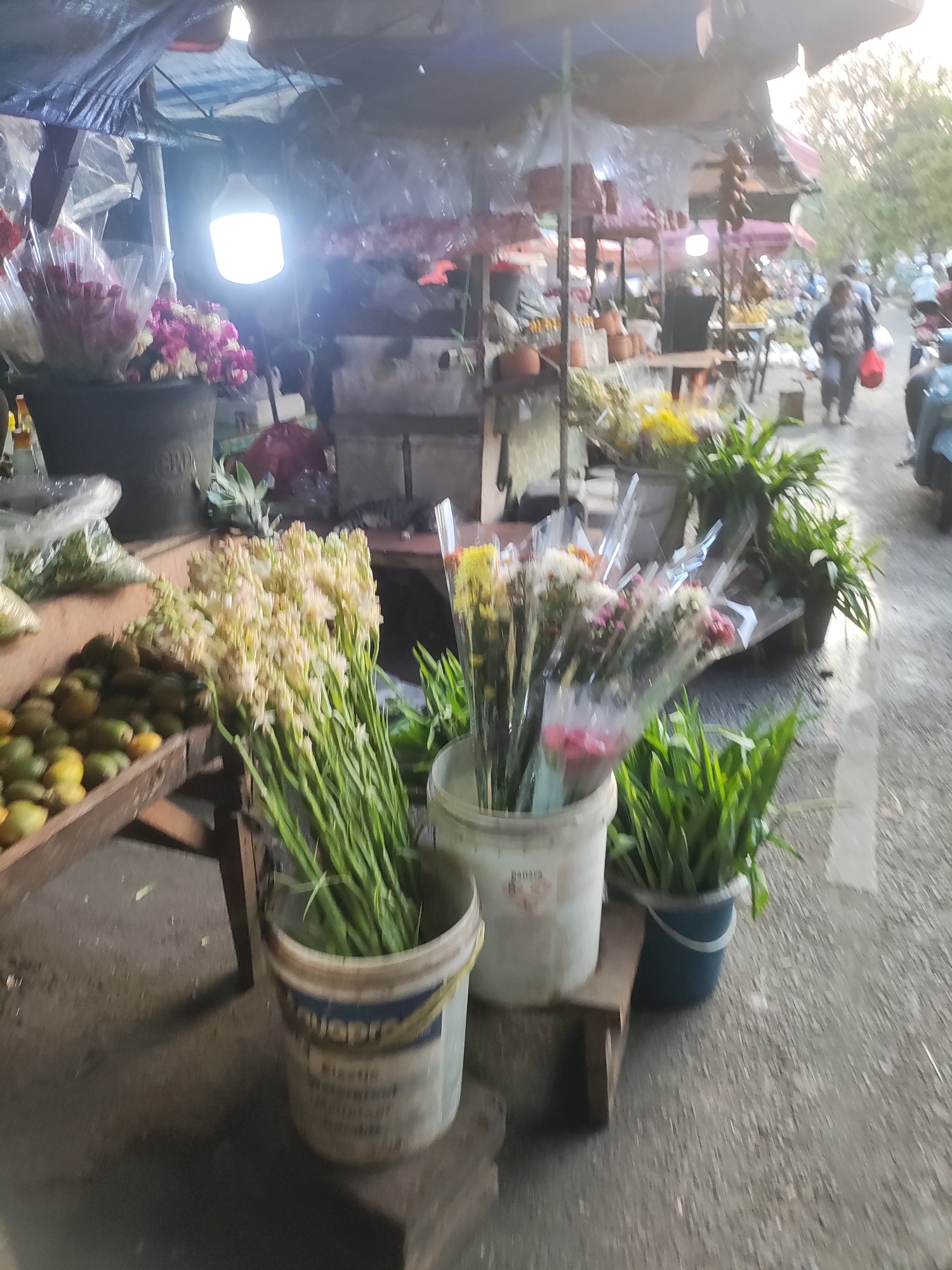 The image depicts an outdoor flower stall at a street market in East Jakarta, Indonesia. The scene is cluttered with various flower arrangements, some in buckets and others wrapped in plastic. In the foreground, a wooden stall displays a collection of green fruits, likely betel nuts. Behind the stall, several buckets hold bunches of white flowers and leafy green plants. The market appears to be bustling, with people walking in the background and a motorbike visible on the right. A bright lightbulb hangs from an overhead tarp, illuminating the stall. The ground is paved and appears wet, suggesting recent rain or a cleaning. The overall atmosphere is that of a typical Southeast Asian street market.