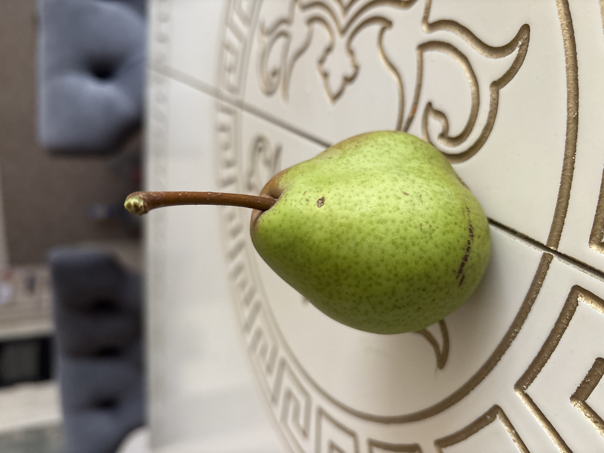 A single, green pear with a brown stem sits on a cream-colored table with a decorative pattern. The pear is positioned in the center of the frame. The table surface is tiled, with gold-colored geometric and ornate patterns. In the blurred background, two out-of-focus gray upholstered chairs are visible, suggesting an indoor setting. The lighting appears to be natural, coming from the front. The image has a shallow depth of field, with the pear in sharp focus and the background softened. There is no discernible location in Georgiyevsk, Russia, indicated in the image.
