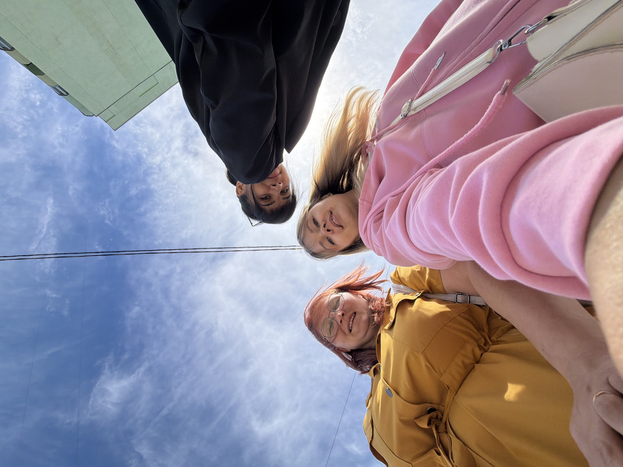 This low-angle photograph captures three young women looking down at the camera against a bright blue sky with scattered white clouds, taken in Chaikovsky, Russia.
On the left, a woman with dark hair and glasses is seen wearing a black hoodie or jacket, looking directly at the camera with a faint smile. In the center, a woman with blonde hair, dressed in a pink hooded sweatshirt and carrying a light-colored shoulder bag, smiles broadly. To her right, another woman with reddish hair and glasses wears a mustard-yellow collared top or dress, also smiling at the camera.
The background is dominated by a clear sky filled with white, wispy clouds. Thin power lines are visible, stretching across the sky, particularly in the middle left portion of the image. In the upper left corner, a section of a light green building with windows is partially visible, suggesting an urban or suburban setting. The bright, natural light indicates it is daytime, likely a sunny morning or afternoon. The overall mood conveyed by the subjects' expressions is cheerful and friendly. There is no visible text in the image.