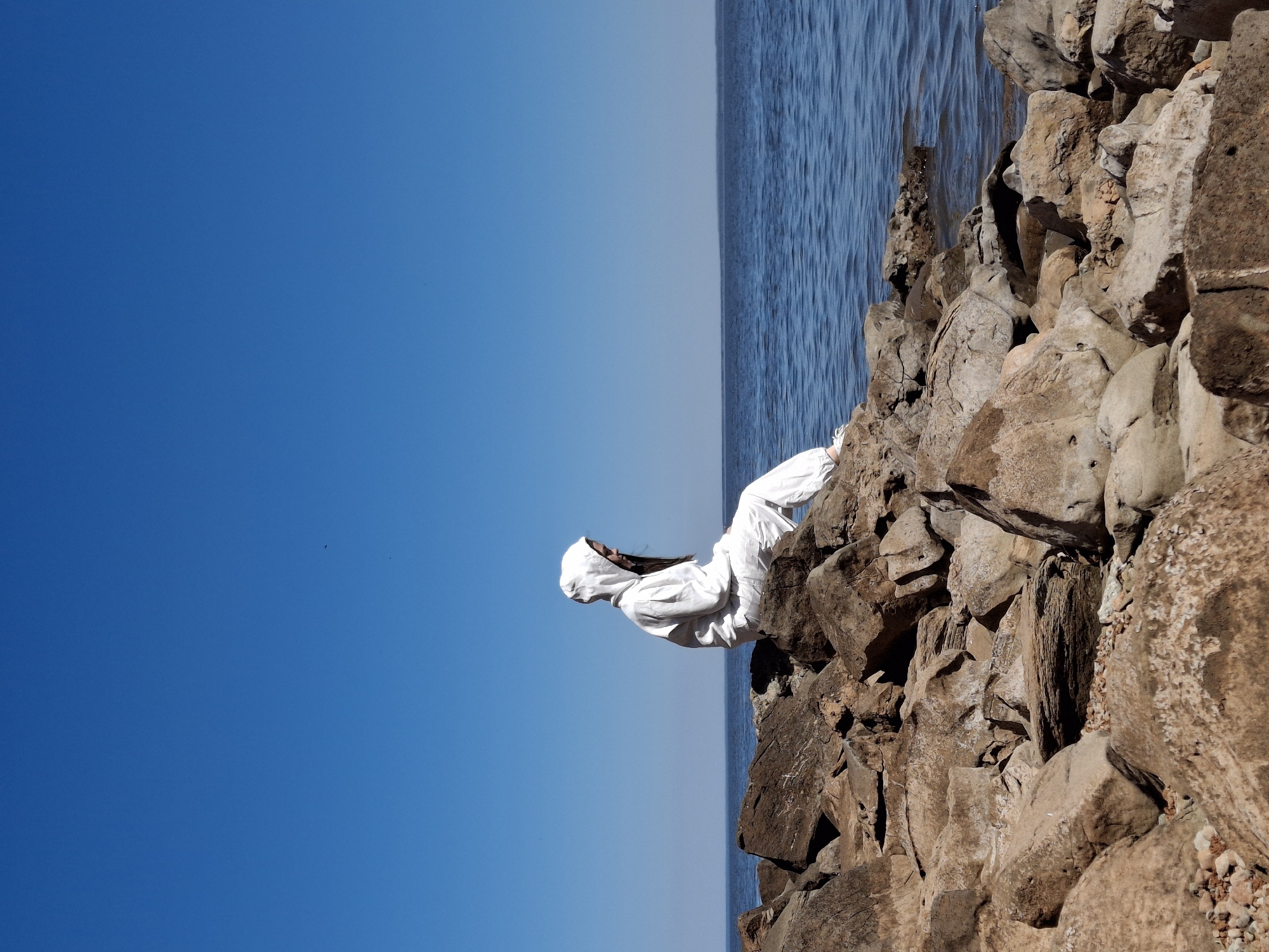 A person in a white hooded garment sits on a rock formation at the edge of the water. The scene is viewed with the image rotated ninety degrees counter-clockwise, orienting the sky to the left side of the frame. The sky is gradient, going from a deeper blue to a lighter blue towards the water. The water is a darker shade of blue. The rock formation consists of large, irregularly shaped stones that appear to be brown and tan. The person is perched on these rocks, facing the water. The surrounding environment appears to be coastal. No precise location is indicated or implied in the scene beyond what is explicitly mentioned.