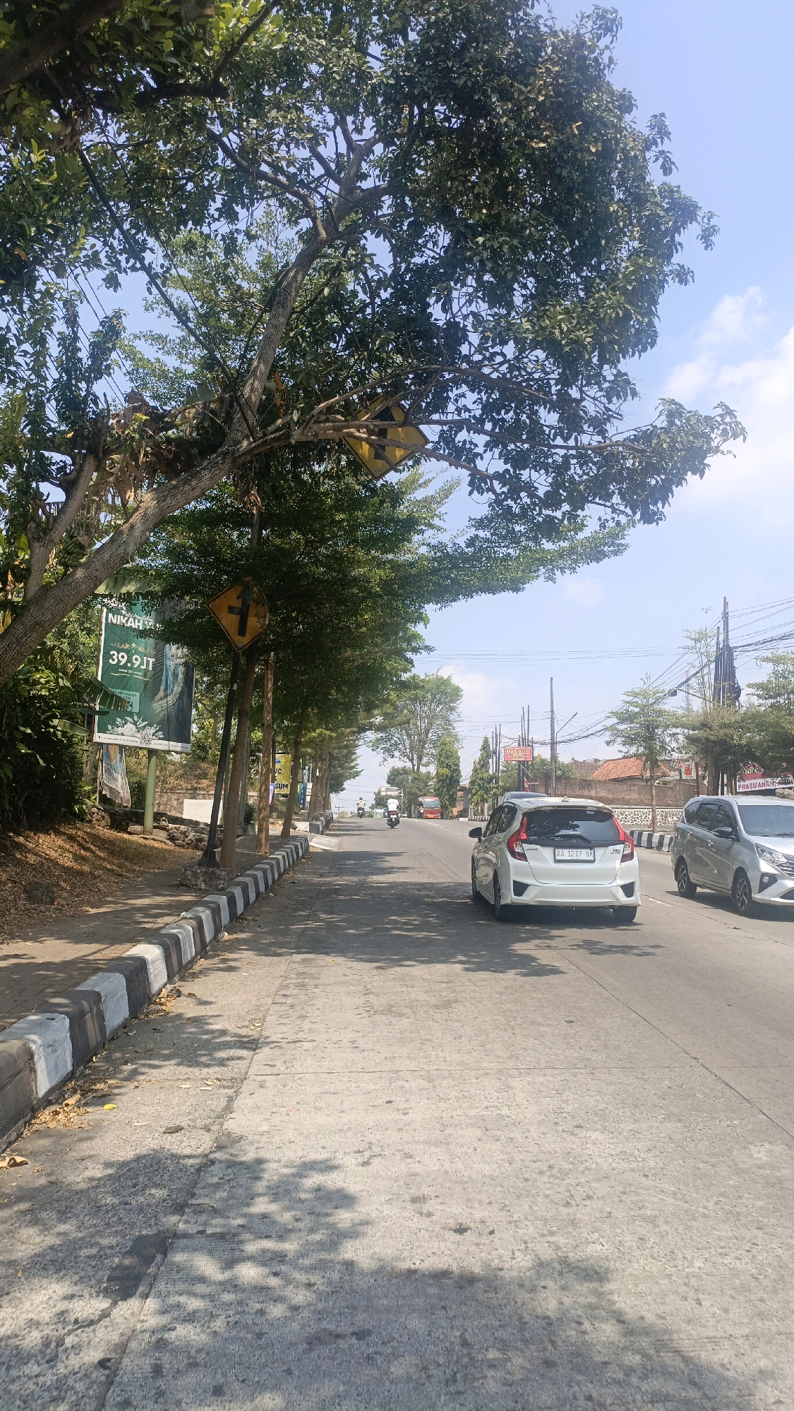 The image depicts a sunny day on a two-lane asphalt road in Salatiga, Indonesia, lined with trees and a sidewalk. A white Honda Jazz is visible in the foreground, followed by a silver MPV. Several traffic signs are present, including a yellow diamond-shaped sign with a black arrow indicating a turn, and a billboard advertising "NIKAH MURAH 39.9JT." The environment appears urban with buildings and power lines in the background. The overall scene suggests a typical street view in a Indonesian city.