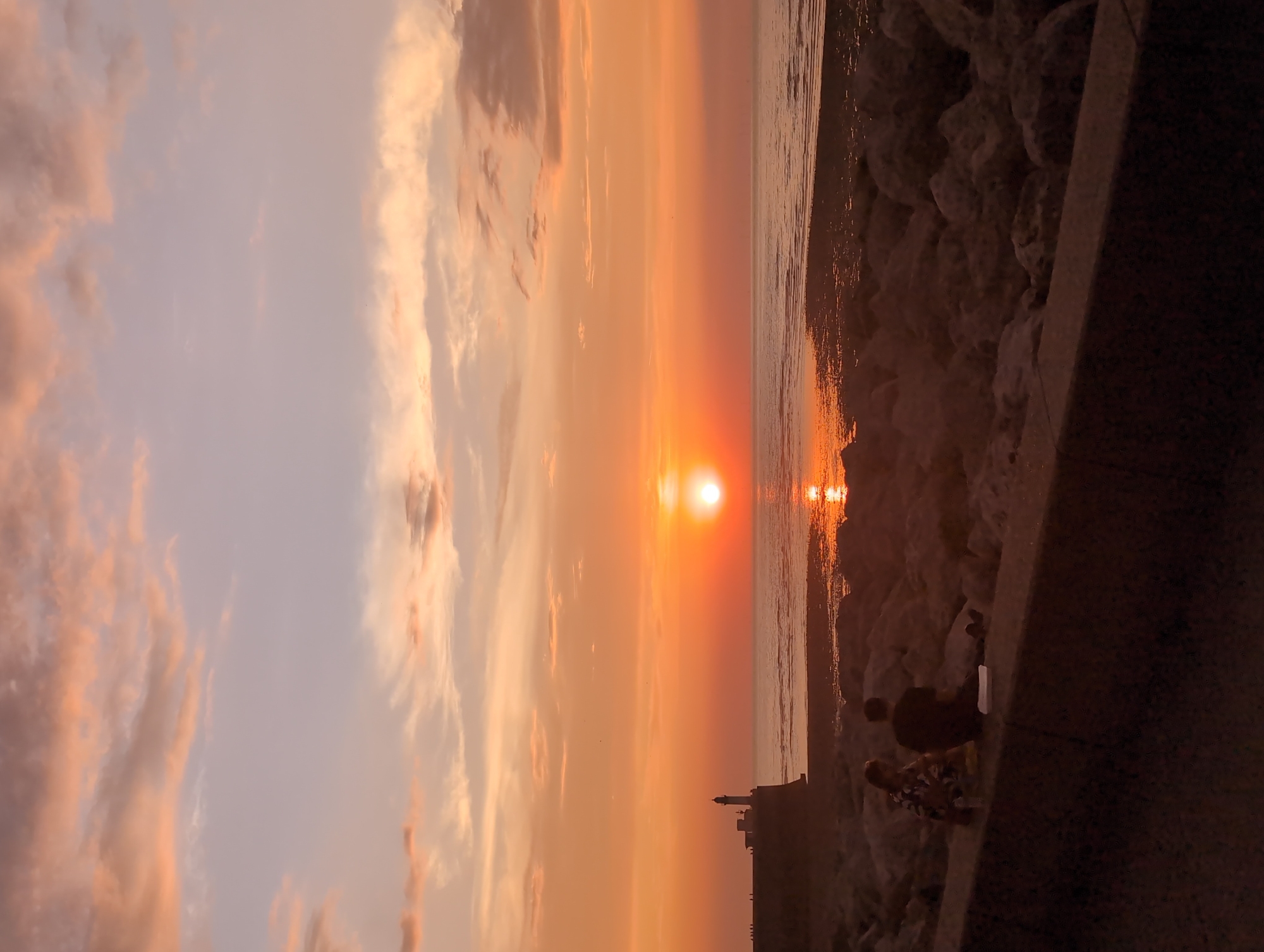 This image captures a sunset over the English Channel at Le Tréport, France. The sky is painted with hues of orange, pink, and purple, with scattered clouds reflecting the warm light of the setting sun. In the distance, a lighthouse stands tall on a stone structure, silhouetted against the vibrant sky. The sea stretches out, with the sun's reflection shimmering on its surface, and a rocky shore is visible in the foreground. Two people are seated on a wall overlooking the sea, appearing to be enjoying the view. One person, dressed in dark clothing, faces away from the camera, while the other, wearing a patterned top, is seated next to them. A few items are placed on the wall in front of them. The overall scene evokes a sense of peace and tranquility.