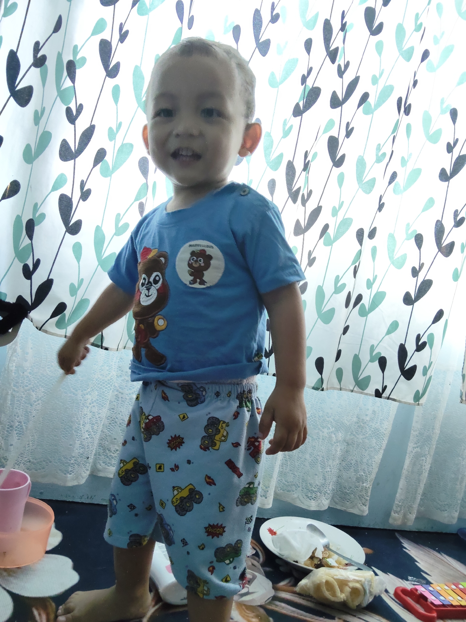 A young child stands in front of a patterned curtain, looking towards the viewer with a slight smile. The child is wearing a blue t-shirt with a bear graphic and light blue shorts adorned with cartoon trucks. The child's right arm is extended, reaching towards the curtain. The environment appears to be indoors, with the patterned curtain suggesting a window or closet. Objects such as a pink cup, a white plate with food, and a colorful toy xylophone are visible on the floor. The lighting suggests it is daytime, with light coming from behind the curtain. The scene is likely a domestic setting in Tanah Grogot, Indonesia.
