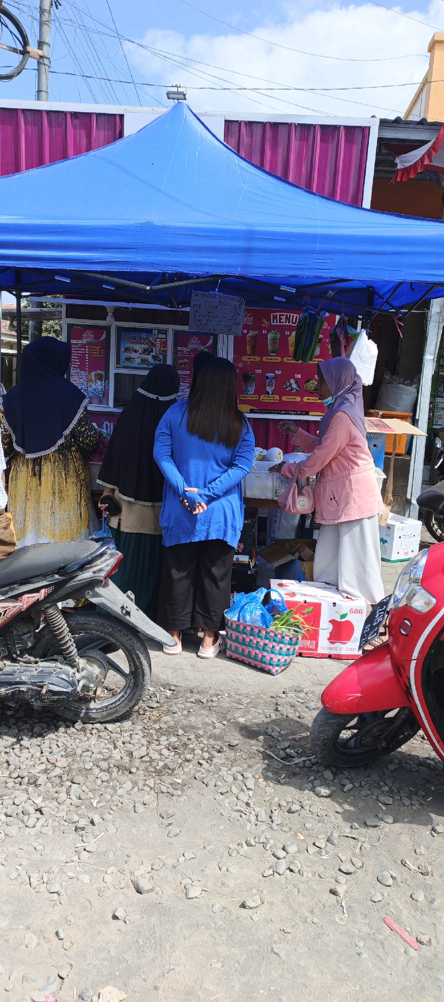 The image depicts an outdoor scene in Puger, Indonesia, featuring a temporary fruit stand with a blue tarp canopy. Several individuals are present, seemingly engaged in a transaction or browsing the offerings. On the left, a person in a yellow and black patterned garment and a dark hijab is visible. Next to them, another individual in a black hijab and light-colored clothing stands. In the center, a person wearing a bright blue long-sleeved shirt and black pants faces away from the camera. To the right, a woman in a pink jacket and white pants, also wearing a headscarf, appears to be interacting with the stand.

The stand itself displays a menu board with various fruit names and prices. Behind the counter, there are coolers and packaged goods. In front of the stand, a pink and green woven basket contains fresh produce, including what appear to be green beans or similar vegetables, and several apples are visible. Two motorcycles are parked near the stand; one is a dark-colored scooter on the left, and a red scooter is partially visible on the right. The ground is covered in gravel and dirt, suggesting a rustic or informal setting. The background includes a building with a pink corrugated metal facade and a light-colored structure, possibly a house or shop. The sky is partly cloudy.