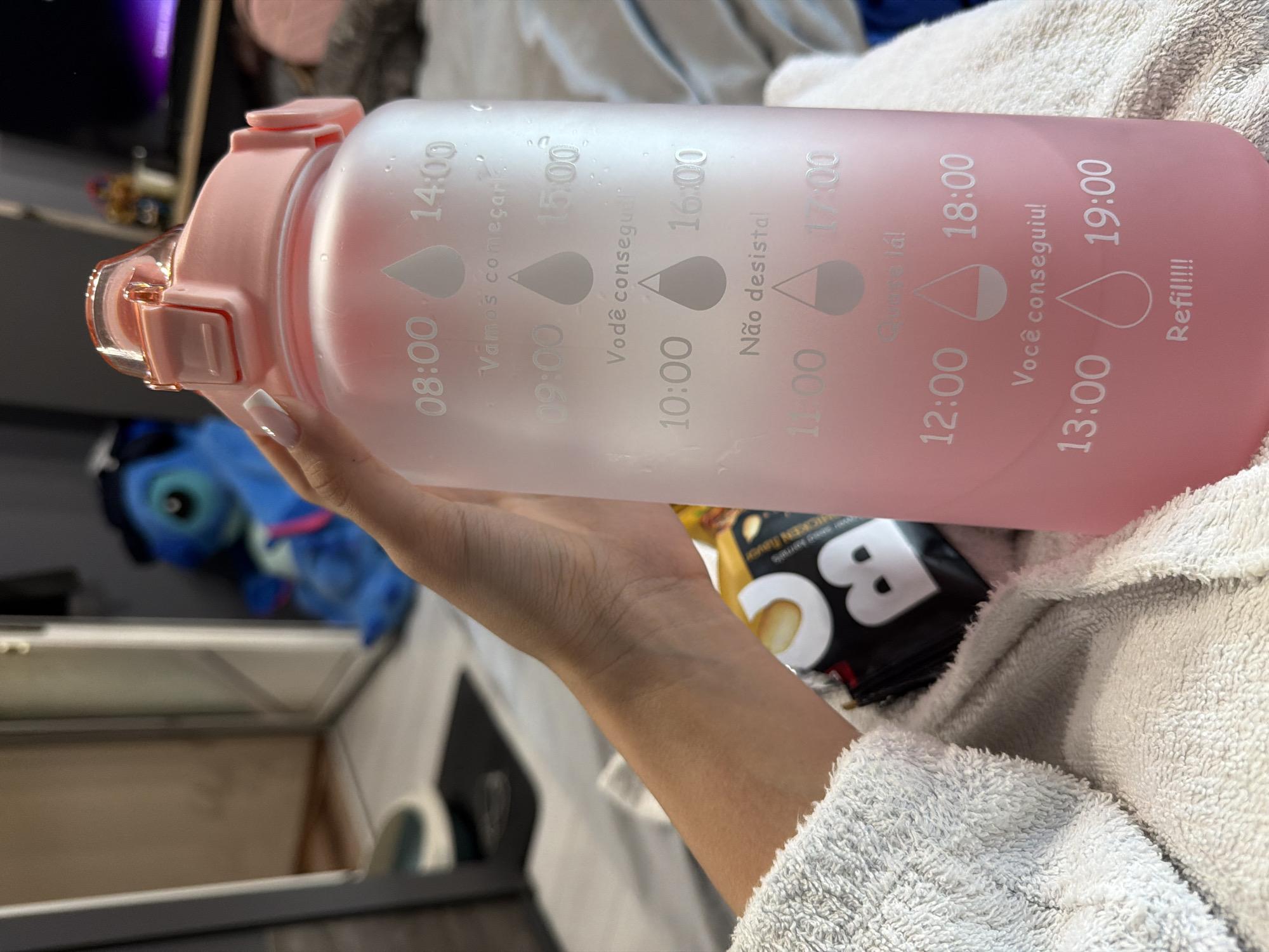 A person's hand with a white-tipped manicure is holding a frosted pink water bottle. The bottle has markings indicating times from 08:00 to 19:00, along with motivational phrases in Portuguese such as "Vamos começar," "Você conseguiu," and "Não desista." In the background, a blue Stitch plush toy is visible, along with what appears to be a television screen and part of a room interior. A white, fluffy towel is draped over the person's arm and the water bottle. A portion of a snack bag with the letter "B" prominently displayed is also in the frame. The scene appears to be indoors, possibly in a bedroom or living area. There is no information in the image to suggest a location in Burgas, Bulgaria.