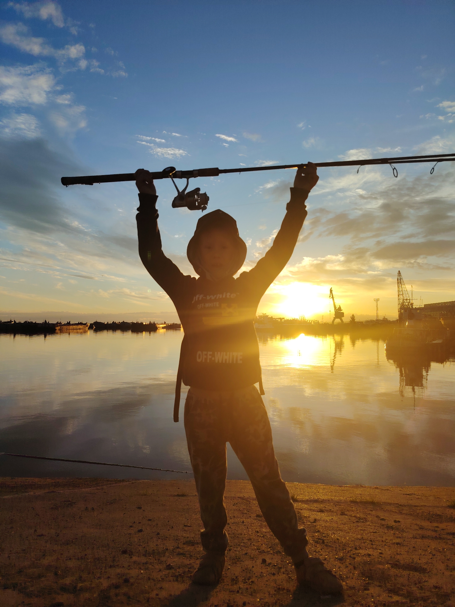 A young person, silhouetted against a sunset, stands on a sandy embankment overlooking a body of water. They are holding a fishing rod aloft with both hands. The person is wearing a hat, a hooded sweatshirt with "OFF-WHITE" text visible, and camouflage pants. The setting sun casts a warm glow across the scene, reflecting on the water's surface. In the background, industrial structures including cranes and ships are visible along the distant shoreline. The sky is a mix of blue and orange hues, with scattered clouds. The overall impression is one of a peaceful, outdoor activity at dusk in a port or industrial area of Chaikovsky, Russia.