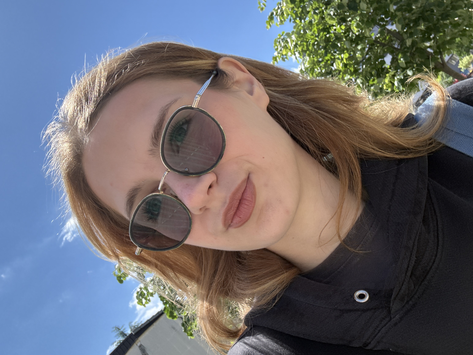 A selfie of a young woman with light brown hair wearing sunglasses and a black hoodie. The background shows a blue sky with some trees and a building. The location is likely outdoors, but it's impossible to pinpoint the exact location in Kragujevac, Serbia from the image alone.