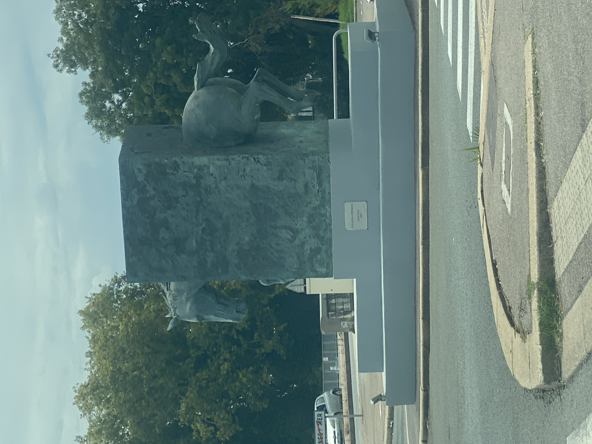 The image shows a bronze sculpture of a horse, with its front half emerging from a rectangular structure and its rear half on the other side. The sculpture is a greenish-blue color and has a textured surface. It is mounted on a grey tiered base. The sculpture is located in Villeneuve-Loubet, France, as indicated by a street sign visible in the background reading "NES MER". The sculpture is situated next to a street with a pedestrian crossing. Trees and cloudy sky are visible in the background.