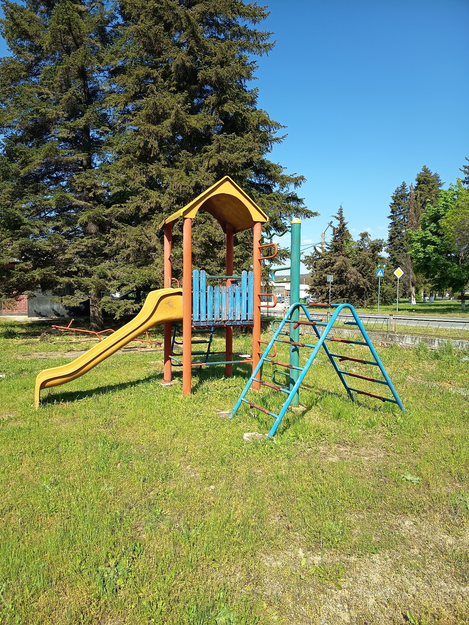 The image shows a children's playground in an urban environment. The playground equipment includes a yellow slide connected to a structure with a yellow roof and blue railings, supported by red posts. A blue and red ladder structure stands nearby. The playground is located on a grassy lawn, with large green trees providing a backdrop. A road runs behind the playground equipment, with traffic signs visible. The sky is clear and blue. There are no people visible in the image. The location appears to be in a park or recreational area in a city.