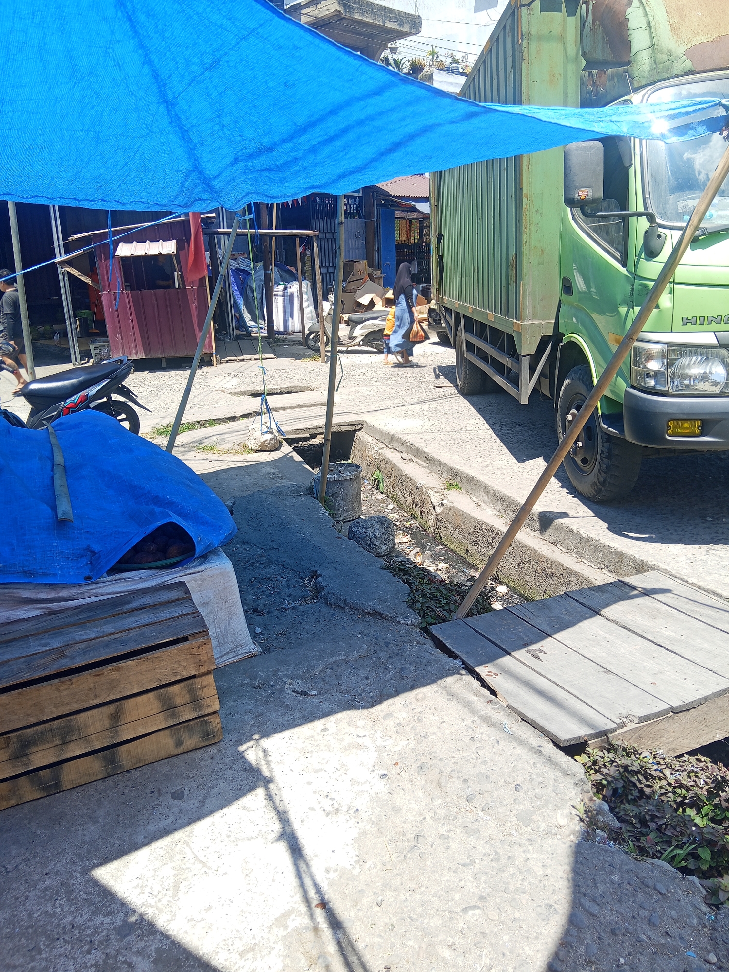 The image depicts an outdoor market scene in Makale, Indonesia. A bright blue tarp is stretched overhead, providing shade. In the foreground, a blue tarp covers a pile of goods on wooden pallets. To the right, a portion of a green truck is visible, its headlights illuminated. A wooden plank bridge crosses a drainage ditch. In the background, a small market stall with a maroon facade is present, along with other structures and a parked motorcycle. Two individuals are visible in the mid-ground, one walking and carrying a bag, the other standing near the stall. The ground is paved with concrete and rough stone. The overall impression is a busy, sunlit market area with goods and vehicles present.