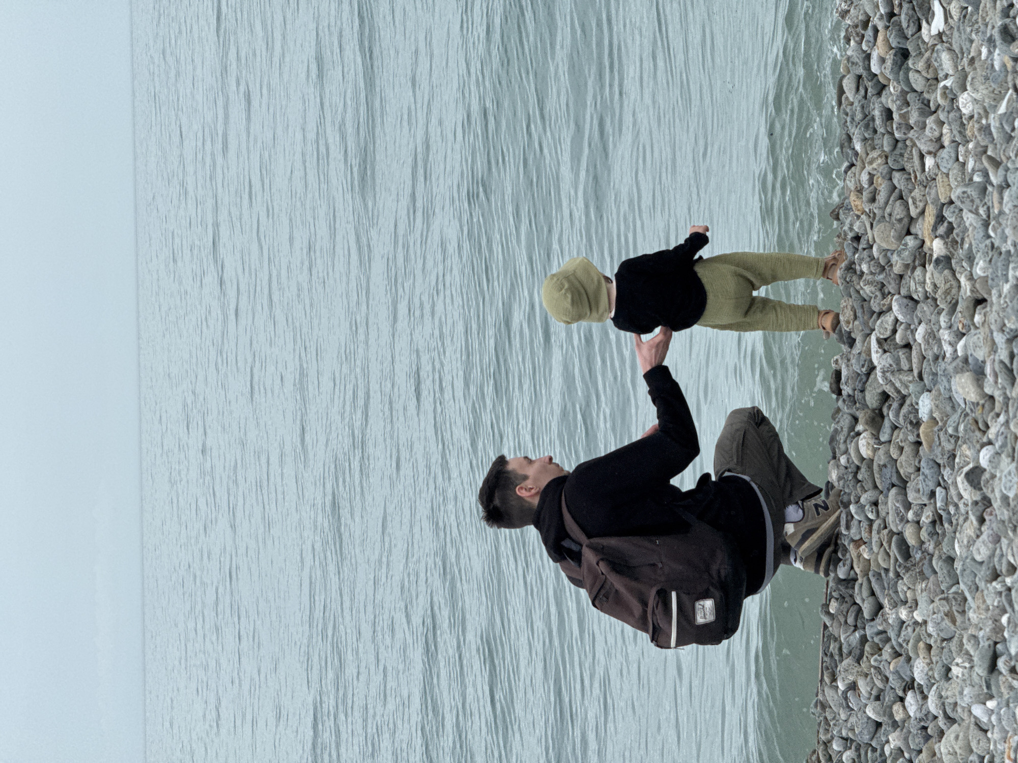 A man with a brown backpack, dressed in a black sweatshirt and gray pants, is sitting on a rocky beach, holding a child, wearing a black sweater, green pants, and a green hat, in front of him. They are both looking at each other. The sea is visible in the background. The photo was taken in the city of Sochi, Russia.
