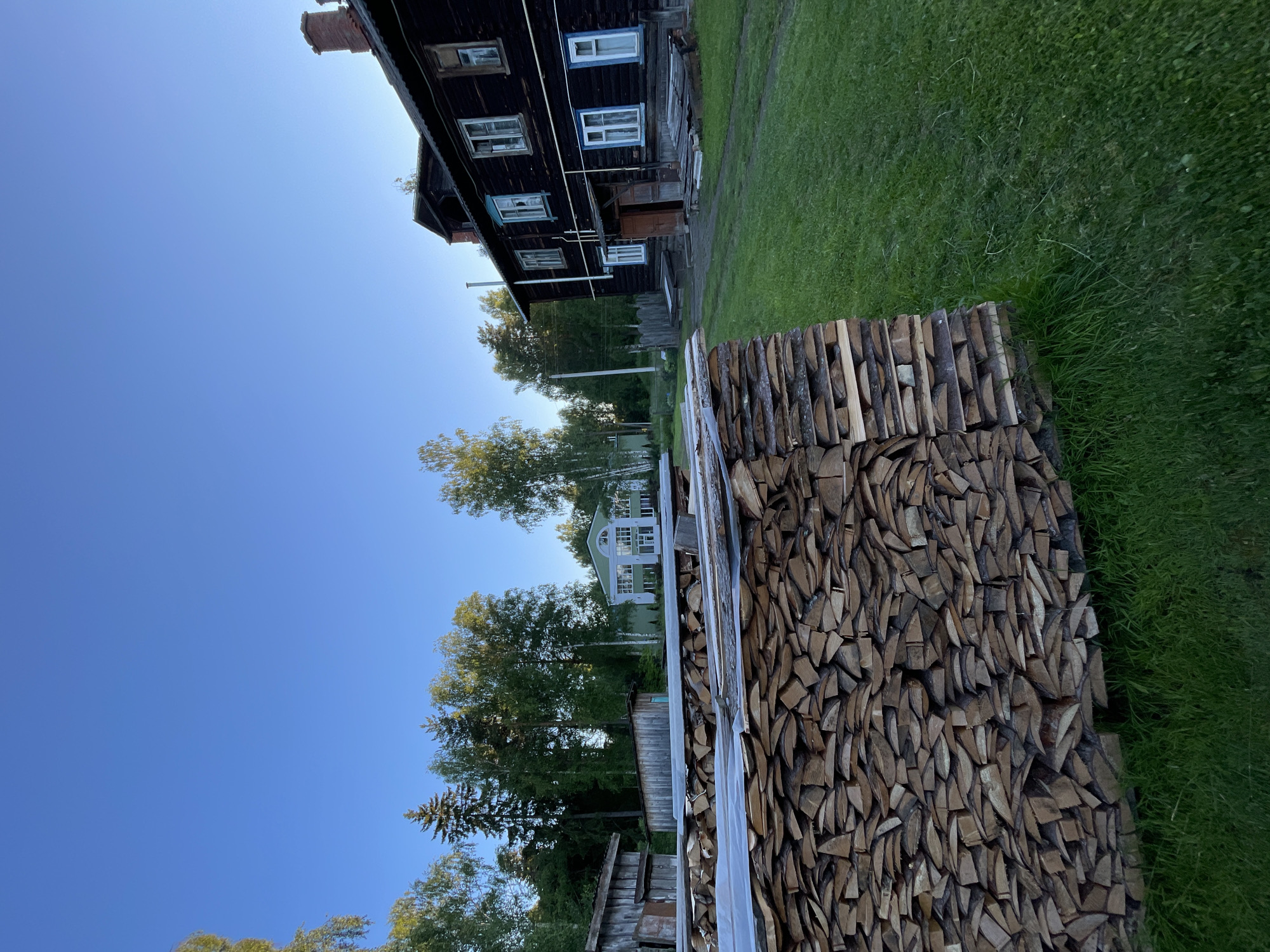 The image depicts a rural scene in Zarechny, Russia, under a clear blue sky, suggesting a daytime setting. The foreground is dominated by a large stack of chopped firewood, neatly arranged and covered with a translucent material. To the left, a weathered, dark wooden house stands, its facade featuring several windows, some with blue trim. Beyond the house, a verdant lawn stretches out, bordered by trees. In the middle distance, a light-colored house with a distinctive arched window on its upper level is visible, nestled amongst lush green foliage. The overall impression is one of a peaceful, rural homestead, possibly in the countryside during summer. There is no visible text in the image.