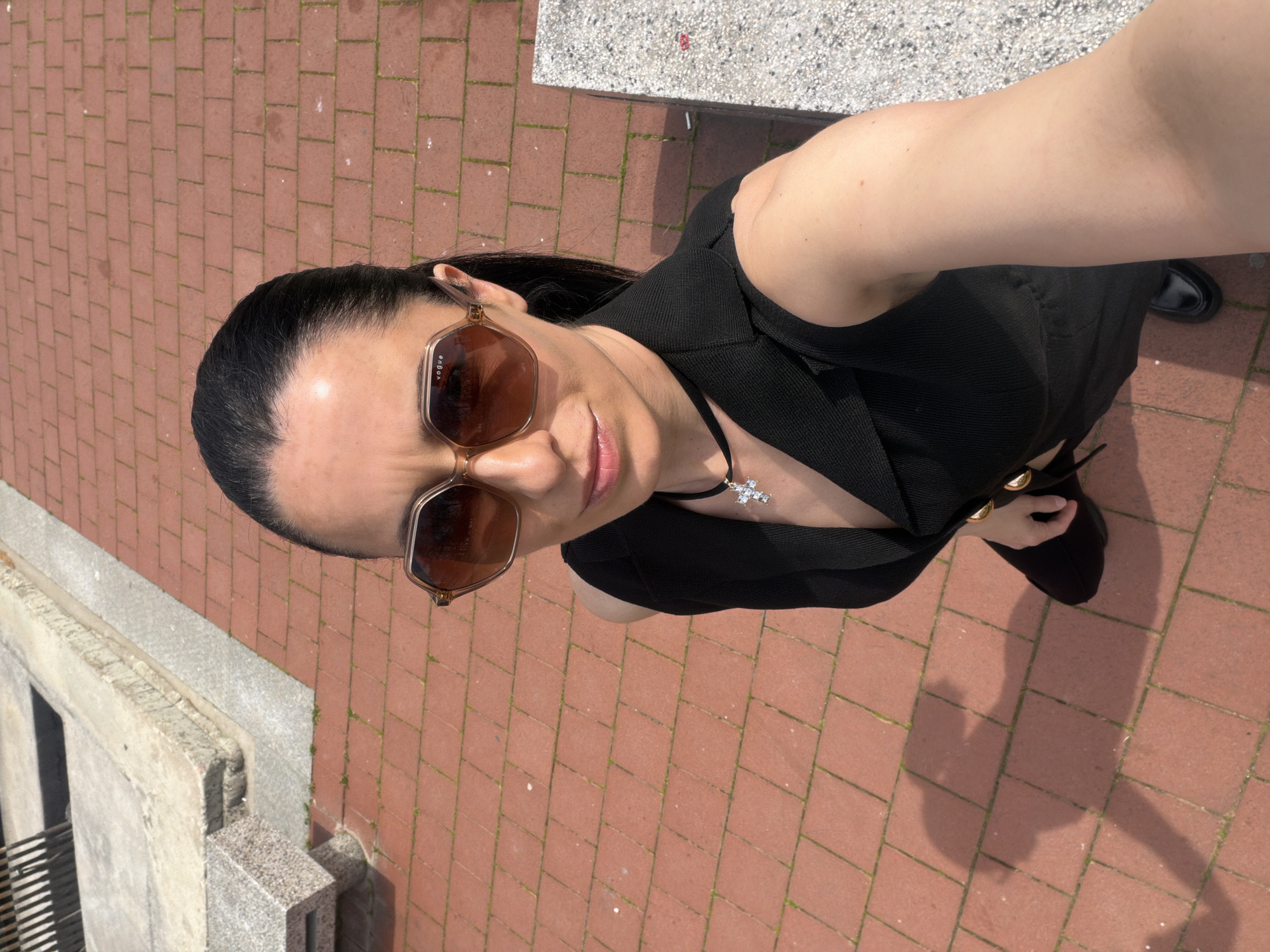 A woman is standing on a red brick pavement. She is wearing a black sleeveless dress, sunglasses, and a necklace with a cross pendant. She is looking up at the camera. The background includes a concrete structure with metal bars and what appears to be a paved area. The location may be in Pleven, Bulgaria.