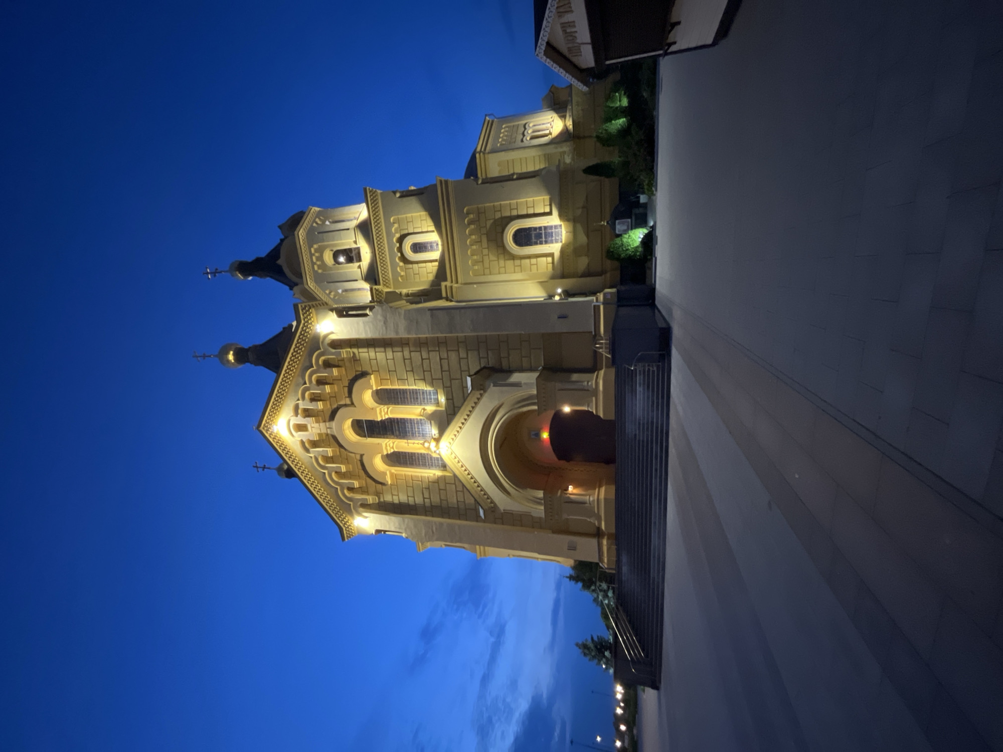 The image depicts the Alexander Nevsky Cathedral in Nizhny Novgorod, Russia, at twilight. The ornate yellow facade of the cathedral is illuminated by warm artificial lights, highlighting its architectural details, including arched windows and domes topped with crosses. The sky is a deep blue, transitioning from a lighter hue near the horizon with faint clouds. A paved pathway leads towards the entrance of the cathedral, which features a wide set of stairs. The pathway is lined with lights, visible in the background. The overall mood is serene and majestic, emphasizing the grandeur of the religious structure. The scene is set in an urban environment, as suggested by the modern pathways and lighting. No people are visible in the frame. There is a partial view of text on a building to the upper right of the image, which appears to be in Cyrillic. The time of day is clearly evening, given the artificial lighting and the dark blue sky.